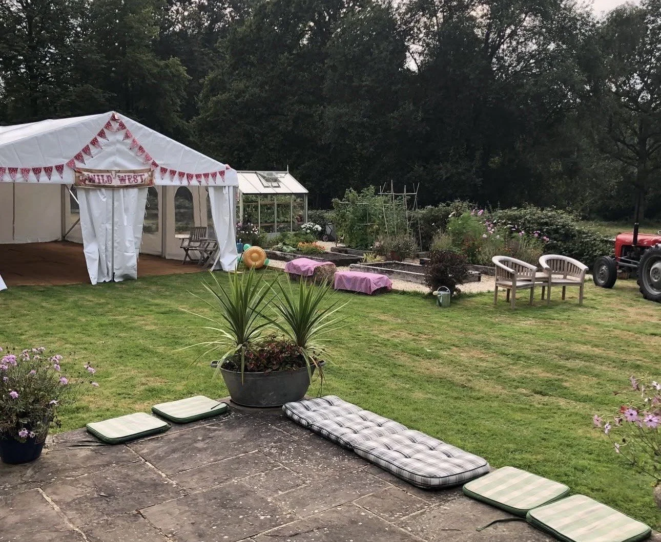 A garden scene with a white tent decorated with pink bunting and a sign that reads 'Wild West.' There are garden beds, a tractor, two white benches, a potted plant, cushions on a stone patio, and lush green trees in the background.