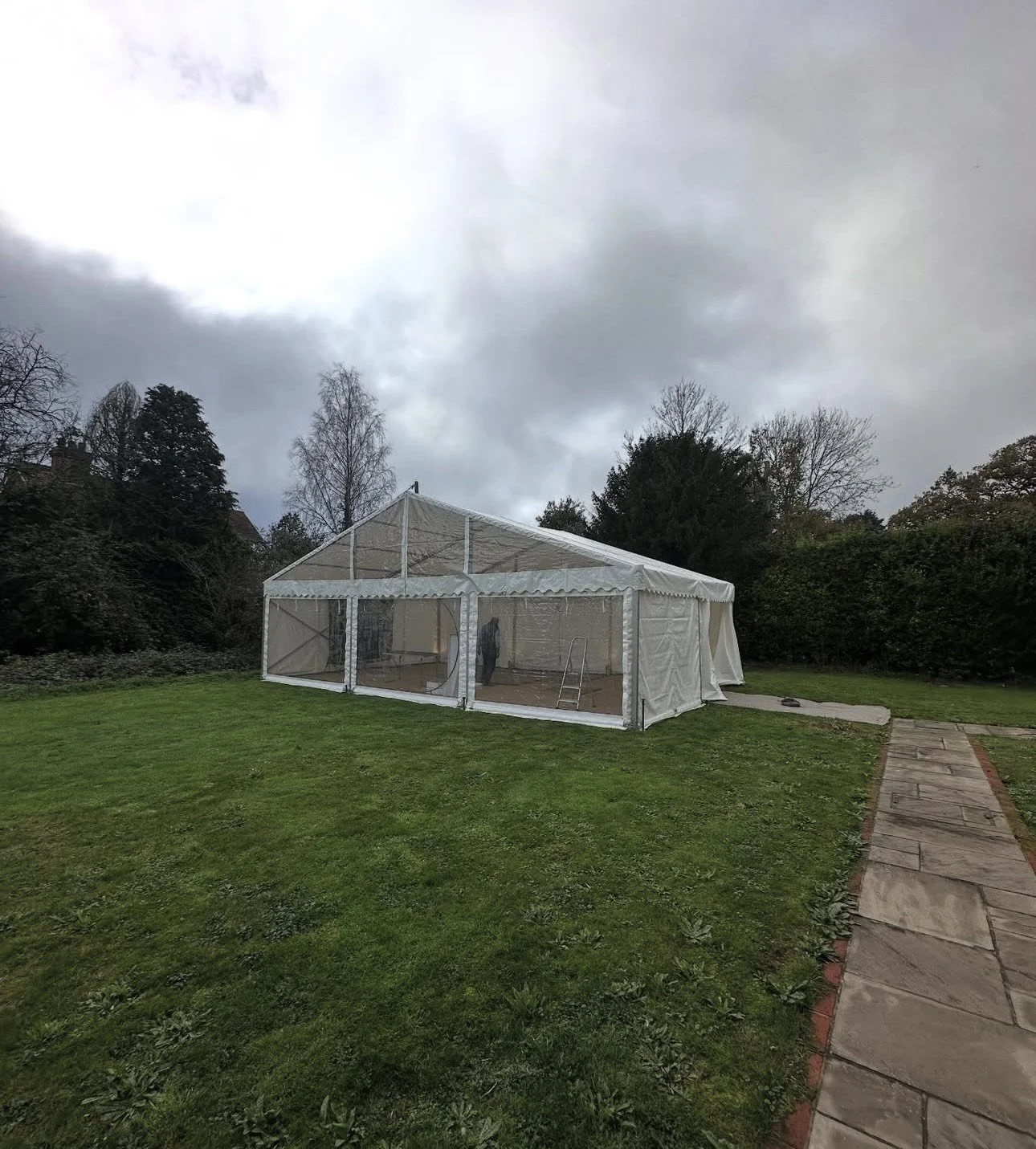 A large white outdoor canopy tent set up on a lawn with trees and bushes in the background, cloudy sky overhead, and a stone pathway leading to the tent.