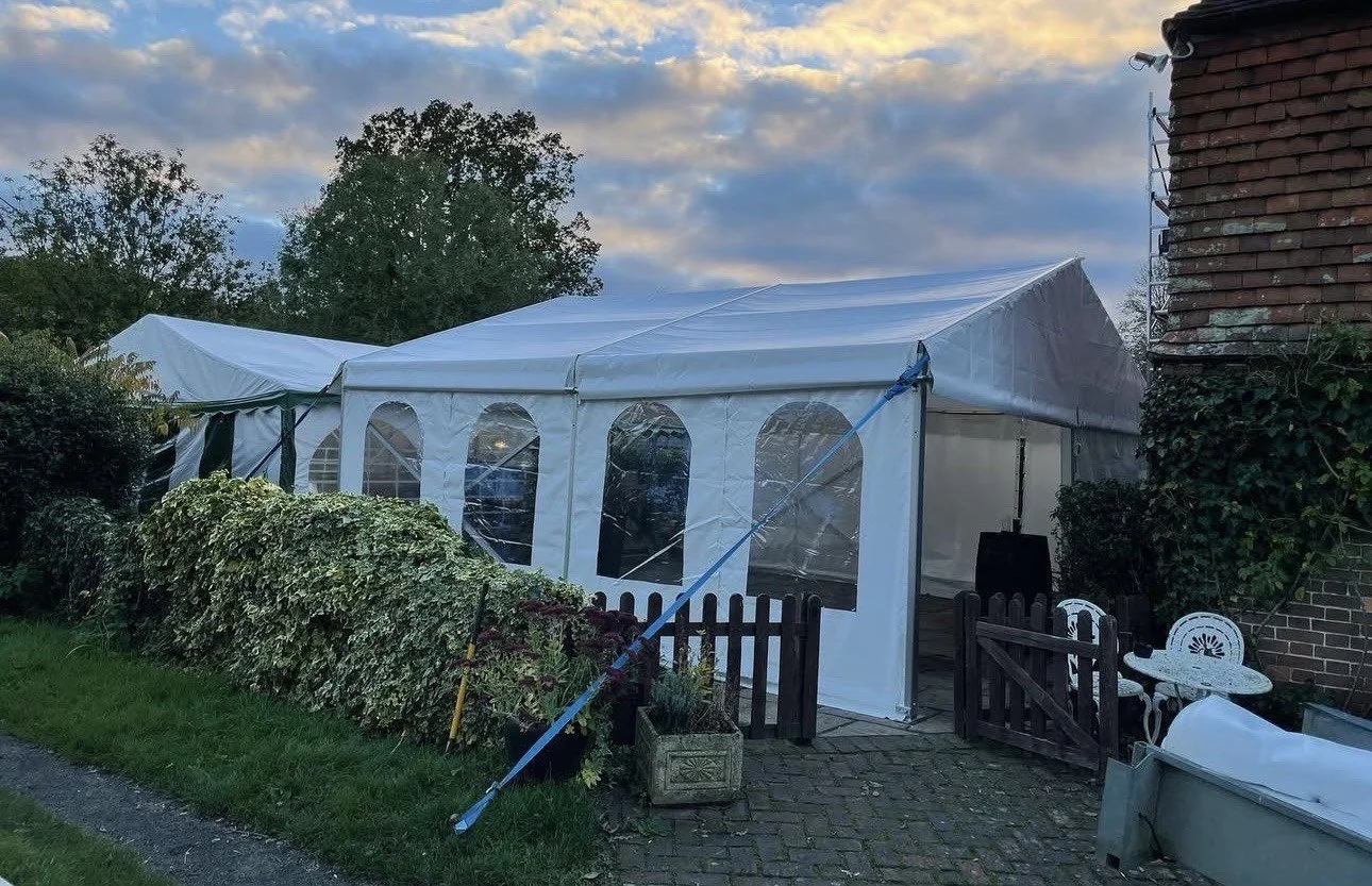 A white outdoor tent set up next to a brick house, with a small garden and patio furniture nearby, under a cloudy sky during sunset.