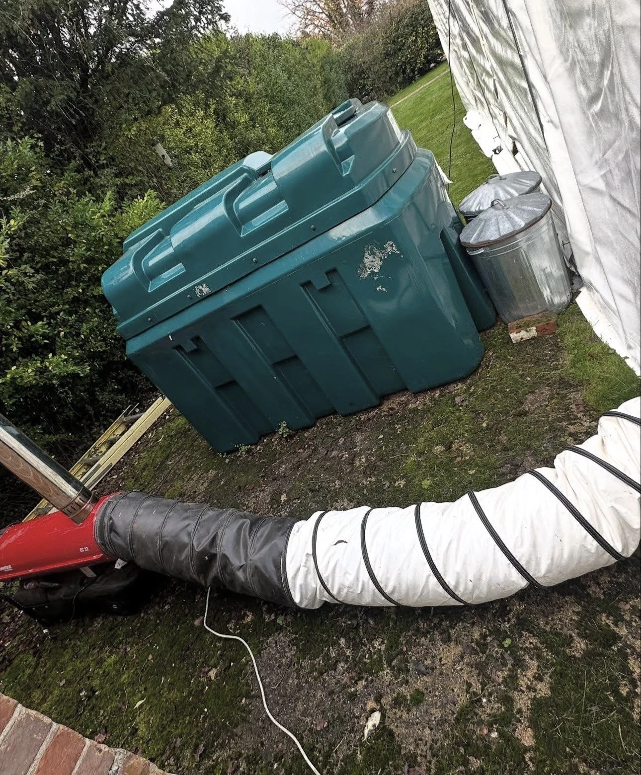 A green trash bin, a silver trash can with a lid, a flexible white and black duct pipe, a red leaf blower, and part of a brick wall on a grassy, dirt surface outdoors.