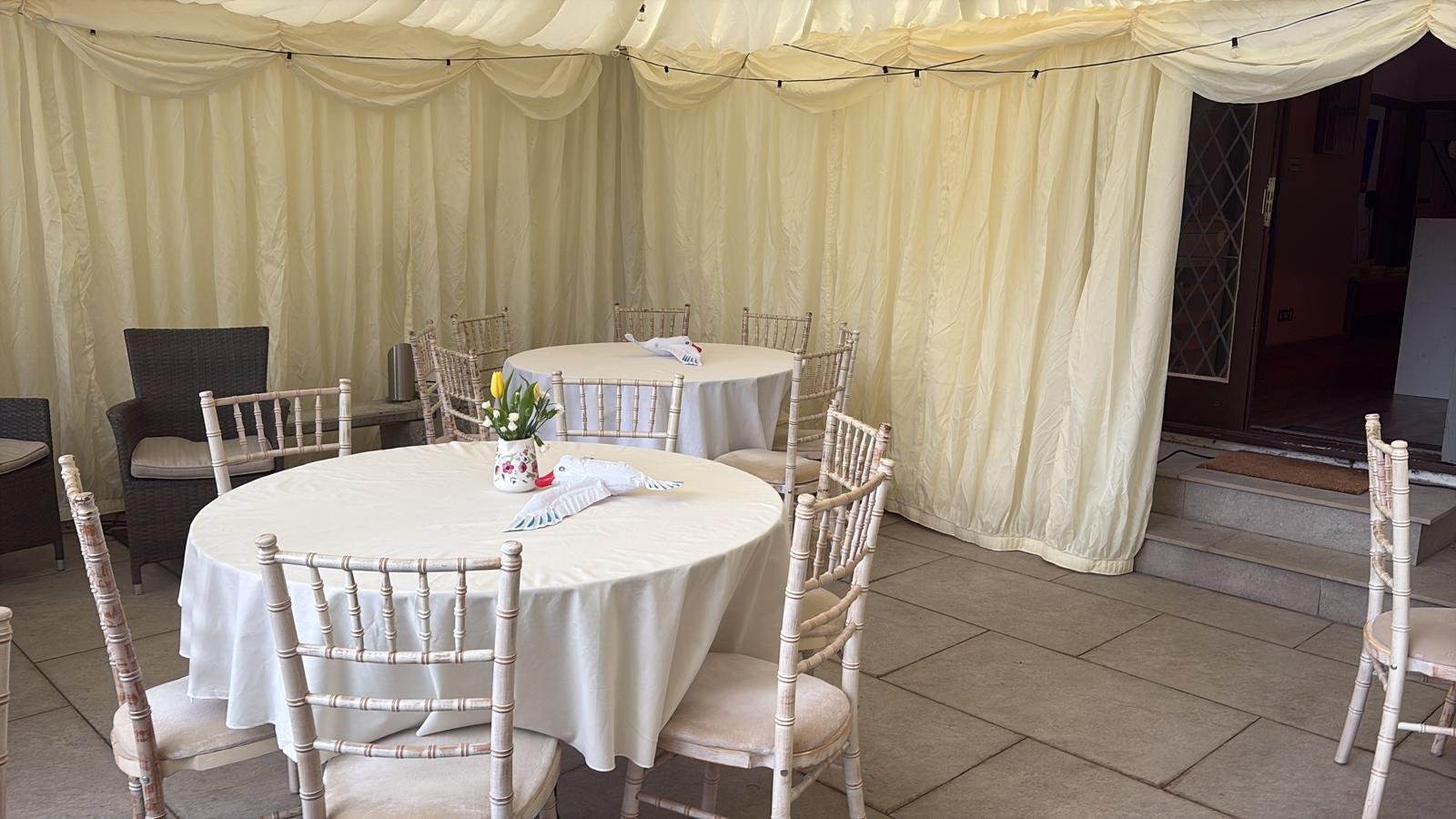 A decorated event space with round tables covered in white tablecloths, each with a vase of yellow tulips, and set against a yellow draped curtain backdrop. There are Chiavari chairs around the tables and dark wicker chairs along the wall.