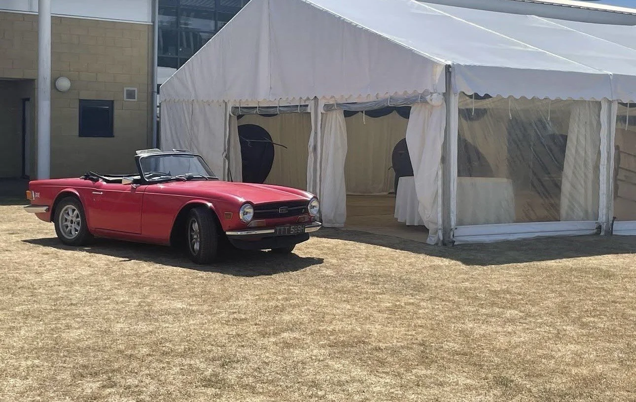 A vintage red convertible car parked on a patchy grass area next to a large white event tent with clear plastic windows.