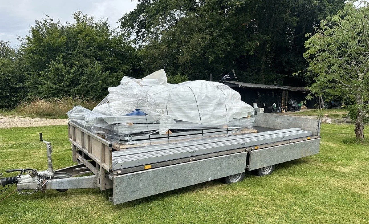 A flatbed utility trailer parked on a grassy area, loaded with large white bags and materials covered partially with plastic wrap, with a background of trees and a small structure.