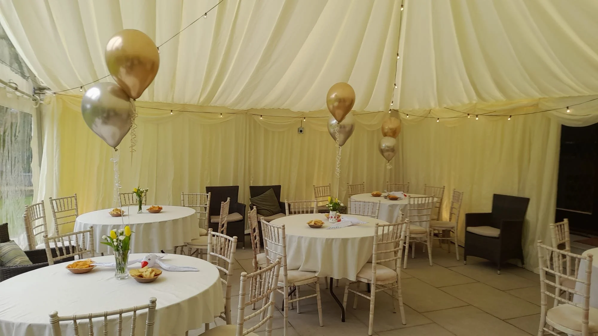 Party tent interior decorated with gold and silver balloons, string lights, round tables with white tablecloths, chairs, and floral centerpieces, set up for a celebration.