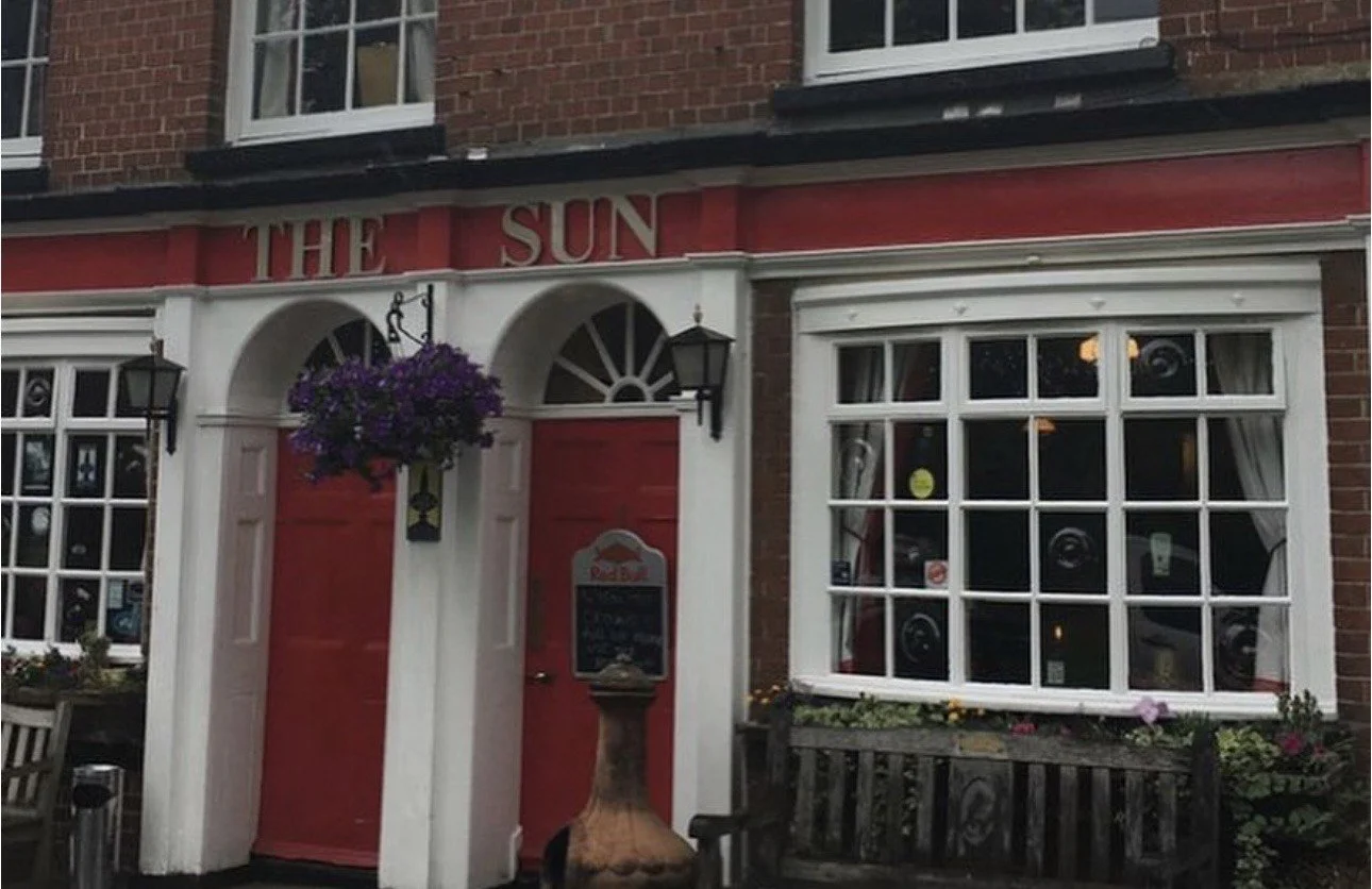 Front view of a brick building with a pub sign 'THE SUN' above a red door, with hanging purple flowers, black lanterns, and a large window with white framing and curtains.