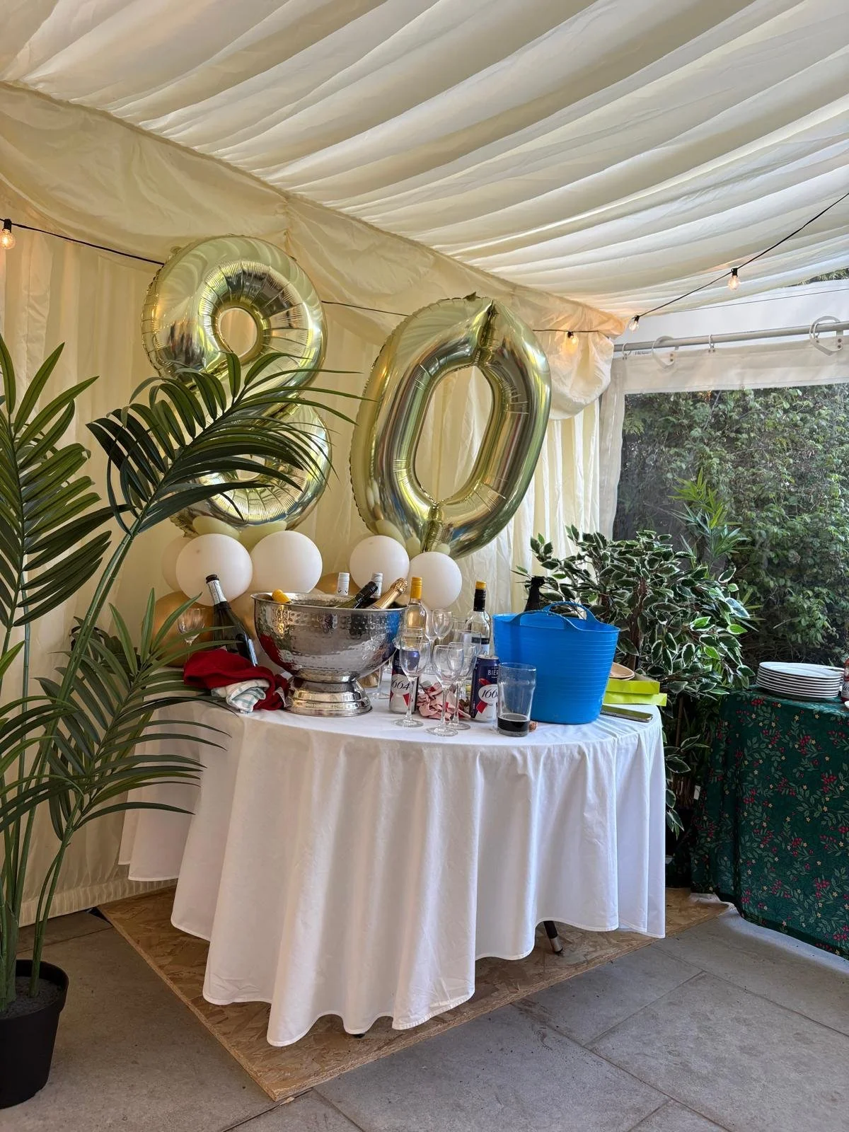 Party table set up inside a tent with large metallic balloons spelling '20', white tablecloth, drinks, and greenery around.