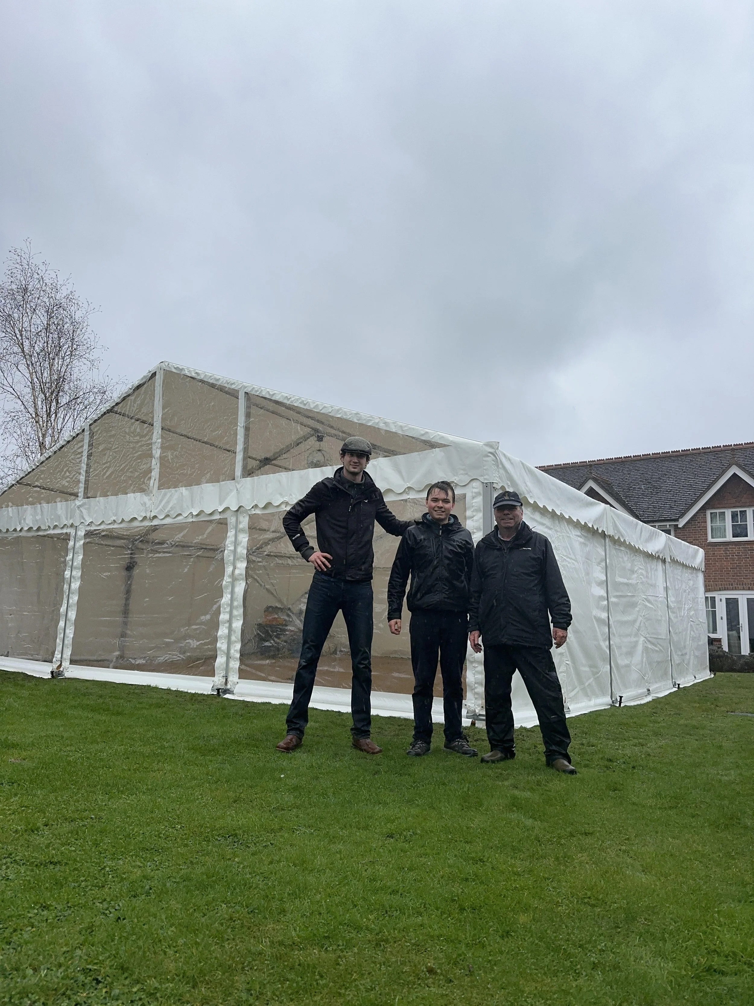 Three men standing in front of a large white event tent on a grassy area on a cloudy day, with residential houses in the background.