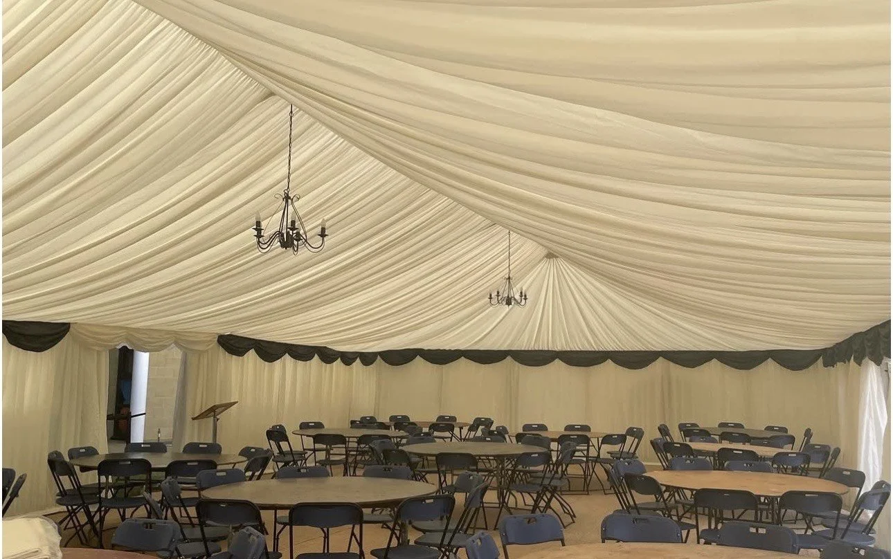 Large event tent interior with beige draped fabric ceiling, black chandeliers, round and rectangular tables with black chairs arranged inside.