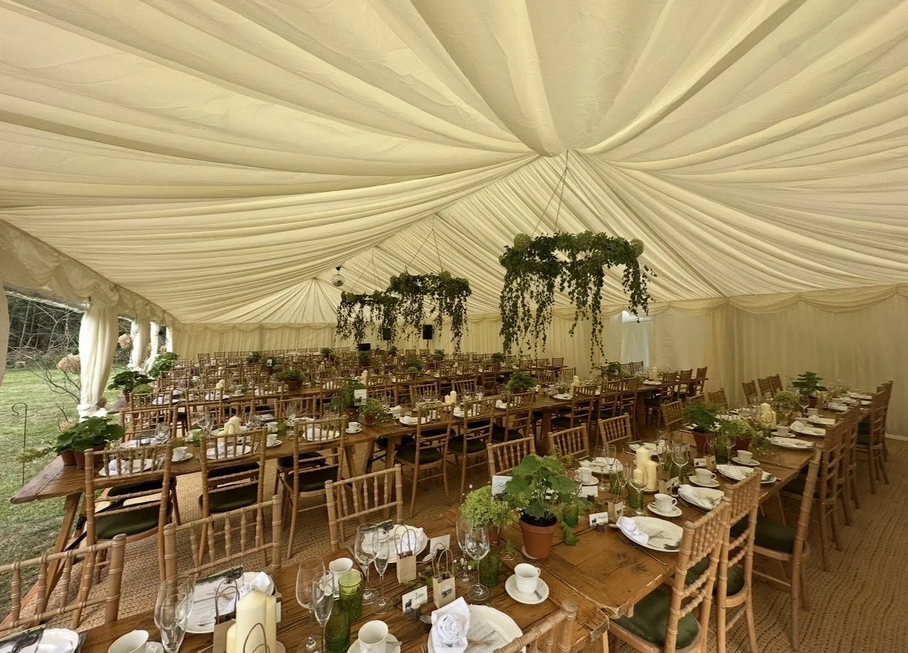 The interior of a large wedding tent decorated with hanging greenery, arranged with long tables set with white plates, glasses, candles, and potted plants for a formal event.