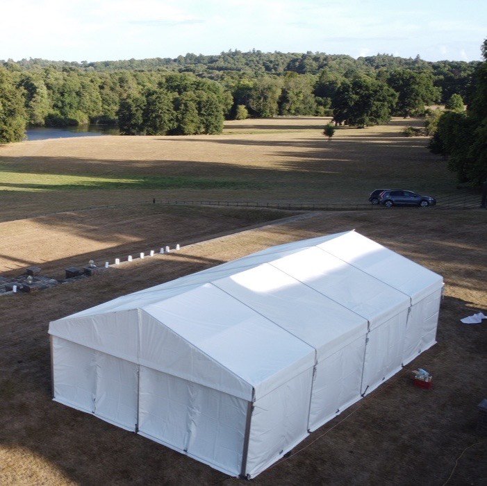 Large white event tent set up on a dry grassy field with a backdrop of trees and rolling hills, and a few parked cars nearby.