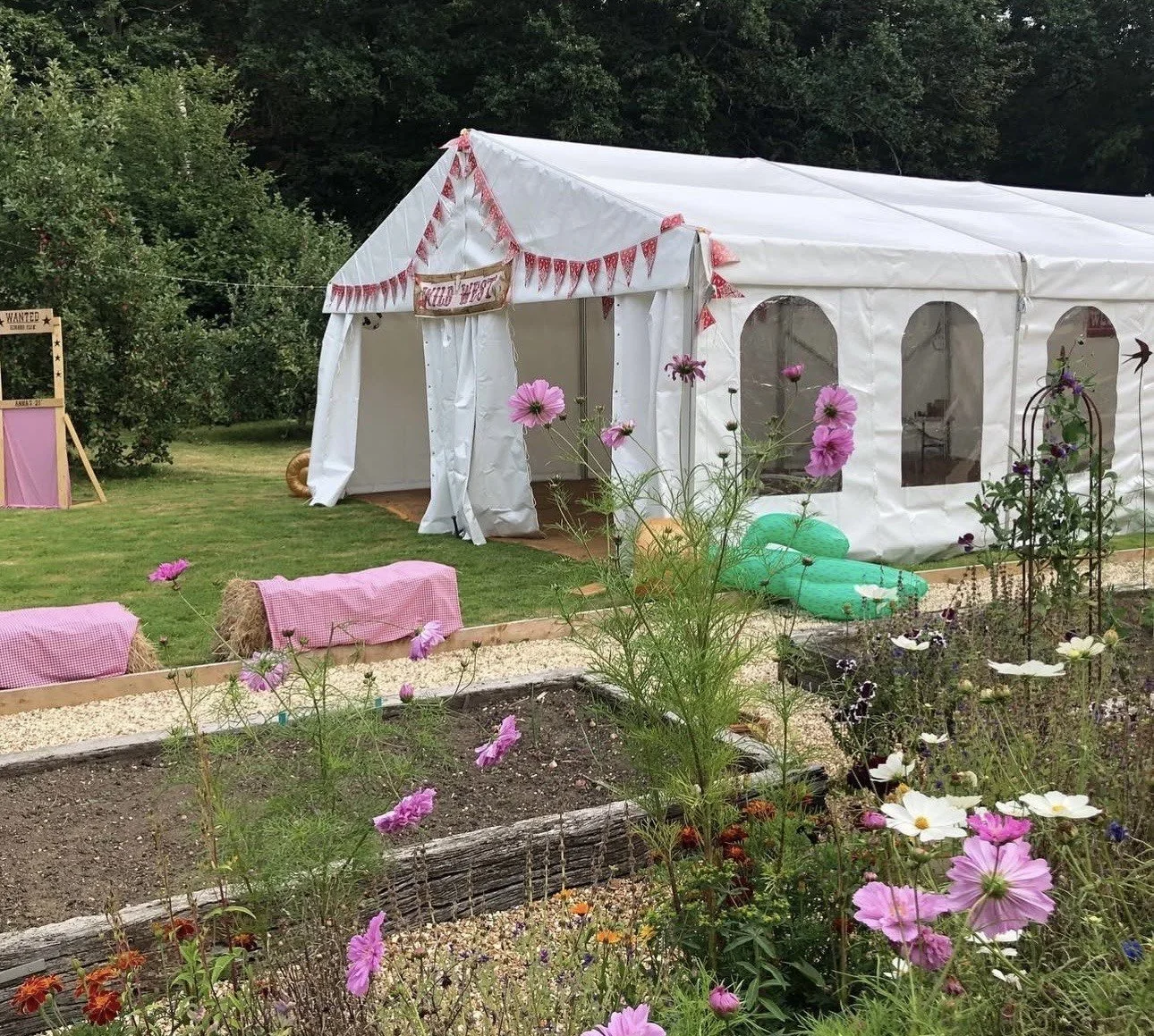 A white event tent with decorations, set up in a garden with colorful flowers and plants, and a pink and white theme for a celebration.
