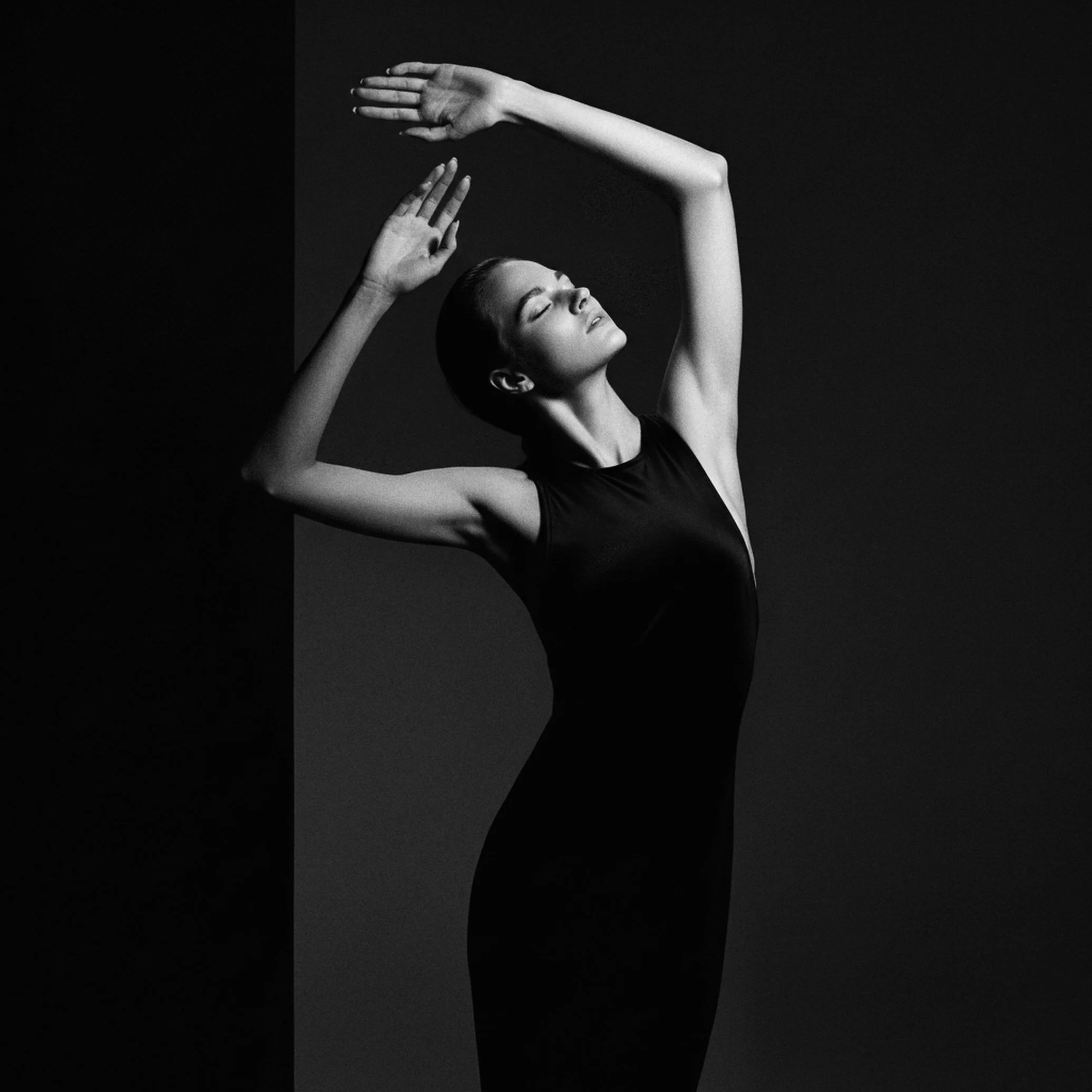 A woman in a black sleeveless dress poses with closed eyes, one arm raised above her head, and the other bent at the elbow with her hand near her face, against a dark background.