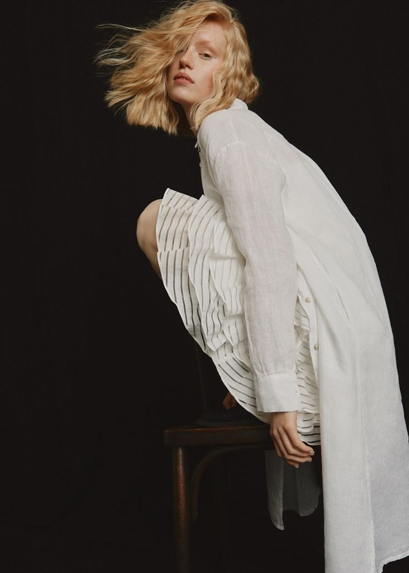 A woman with blonde hair posing against a black background, dressed in a white linen coat and a striped skirt, sitting on a wooden chair with one knee raised.