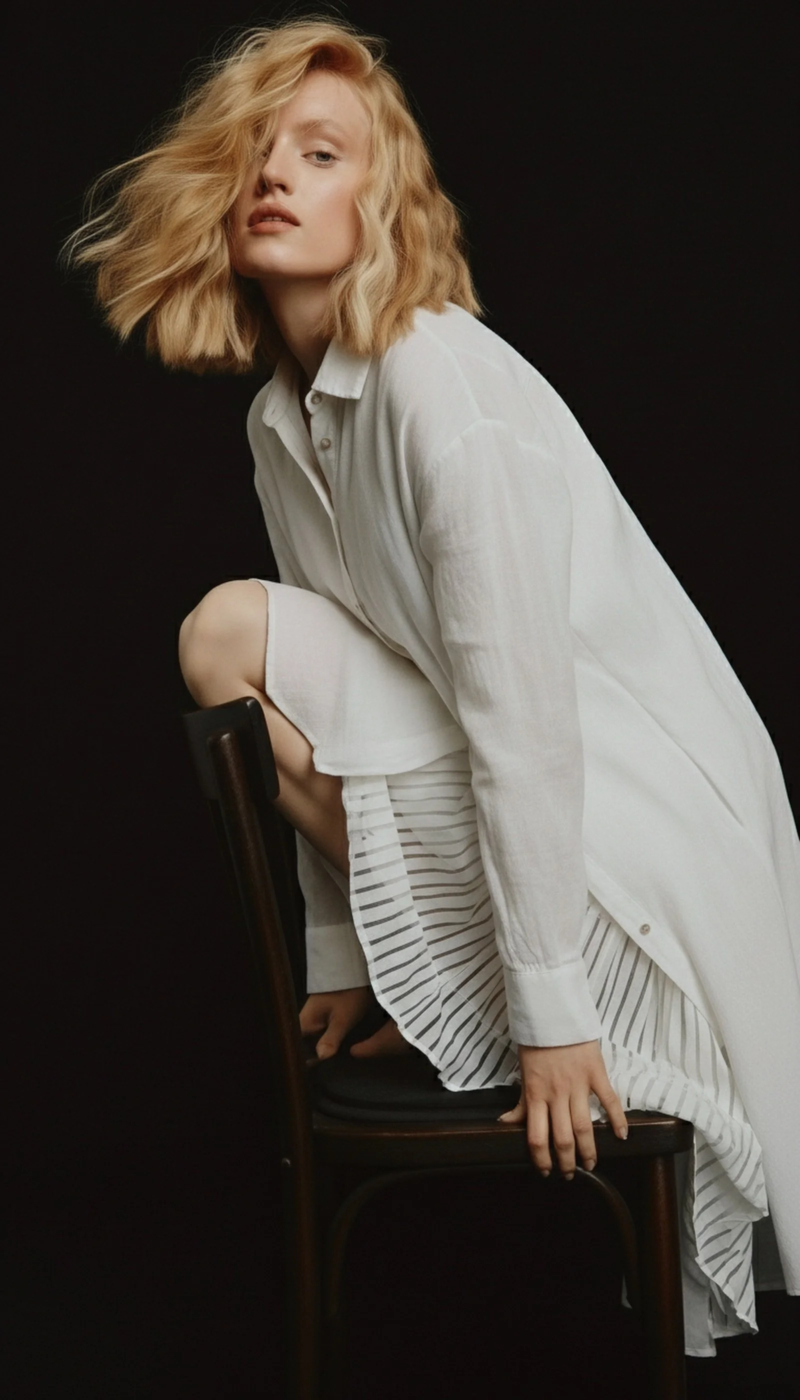 A woman with blonde curly hair wearing a white shirt and skirt, sitting with one knee up on a chair against a dark background.