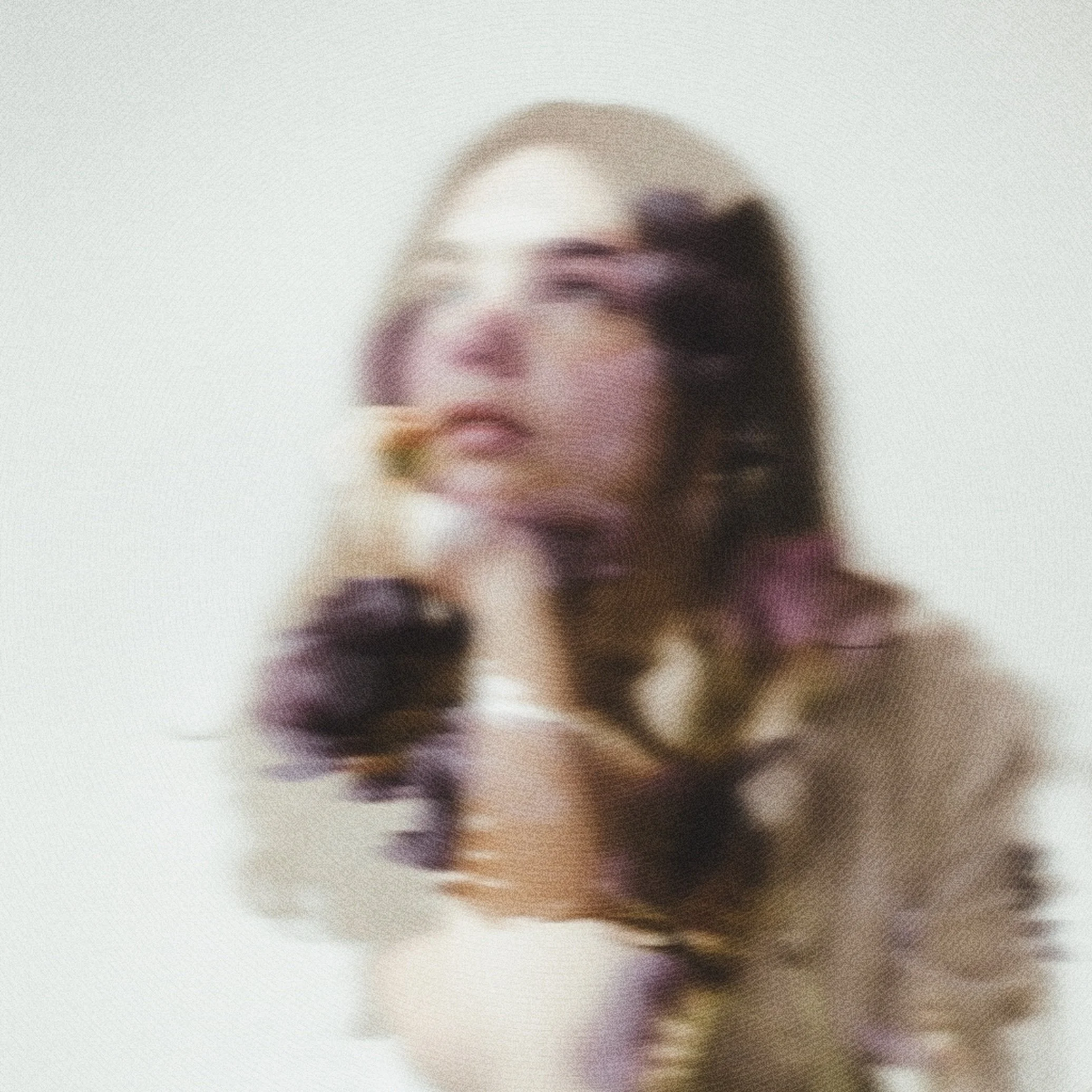 A blurred photo of a woman with dark hair holding a flower near her chin, standing against a plain white background.