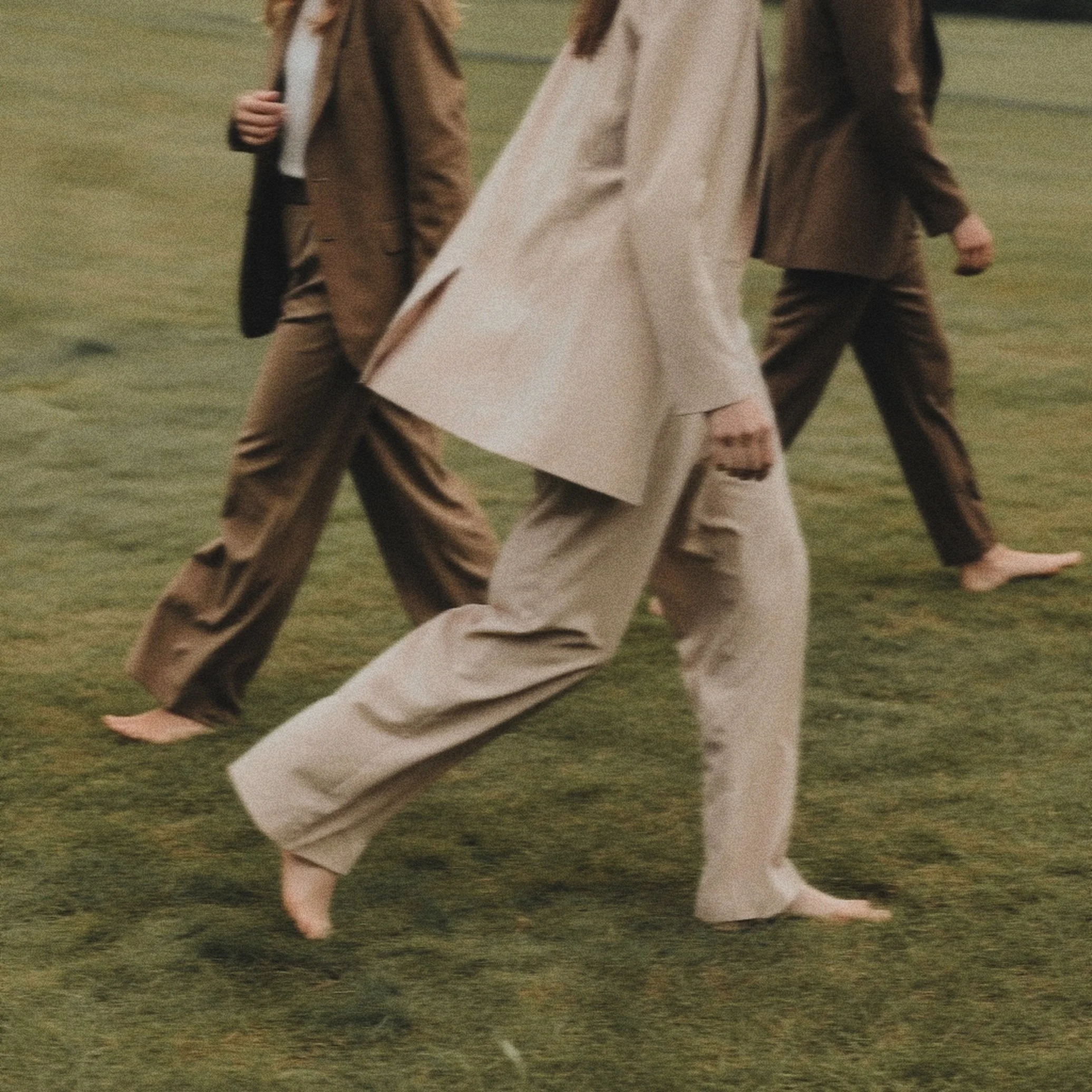 Barefoot individuals walking on a grassy field, dressed in brown and beige suits and coats.