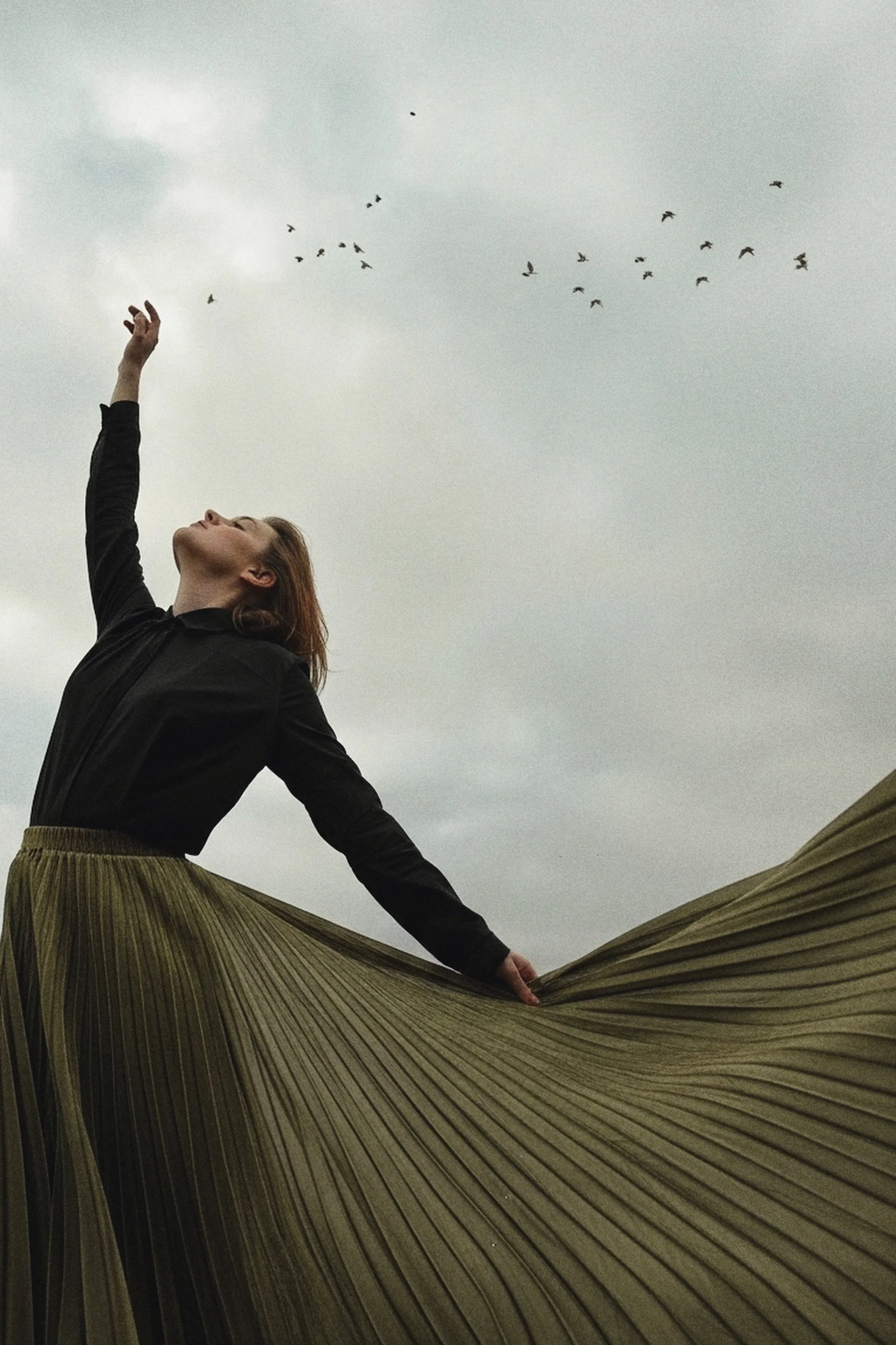 A woman wearing a black jacket and a long, pleated green skirt standing outdoors against a cloudy sky, with her arm raised and head tilted back, while holding her skirt out to the side.