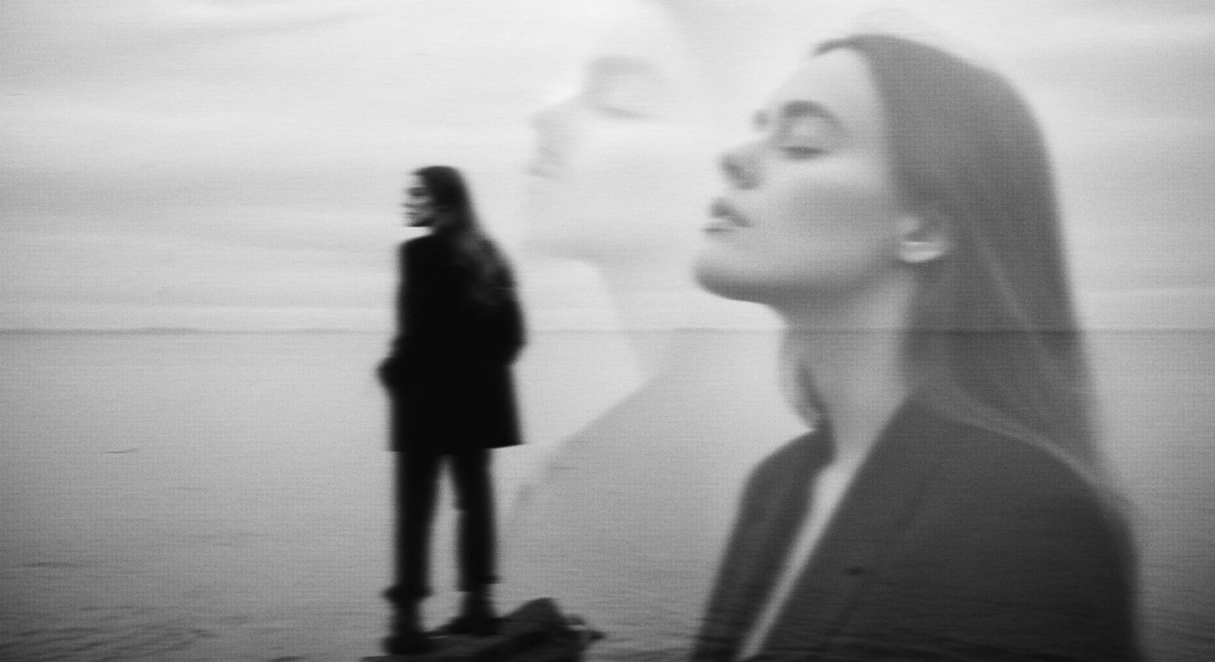 Black and white image of two women, one in the foreground with eyes closed and head slightly tilted, and another in the background standing on a beach, with an overcast sky.