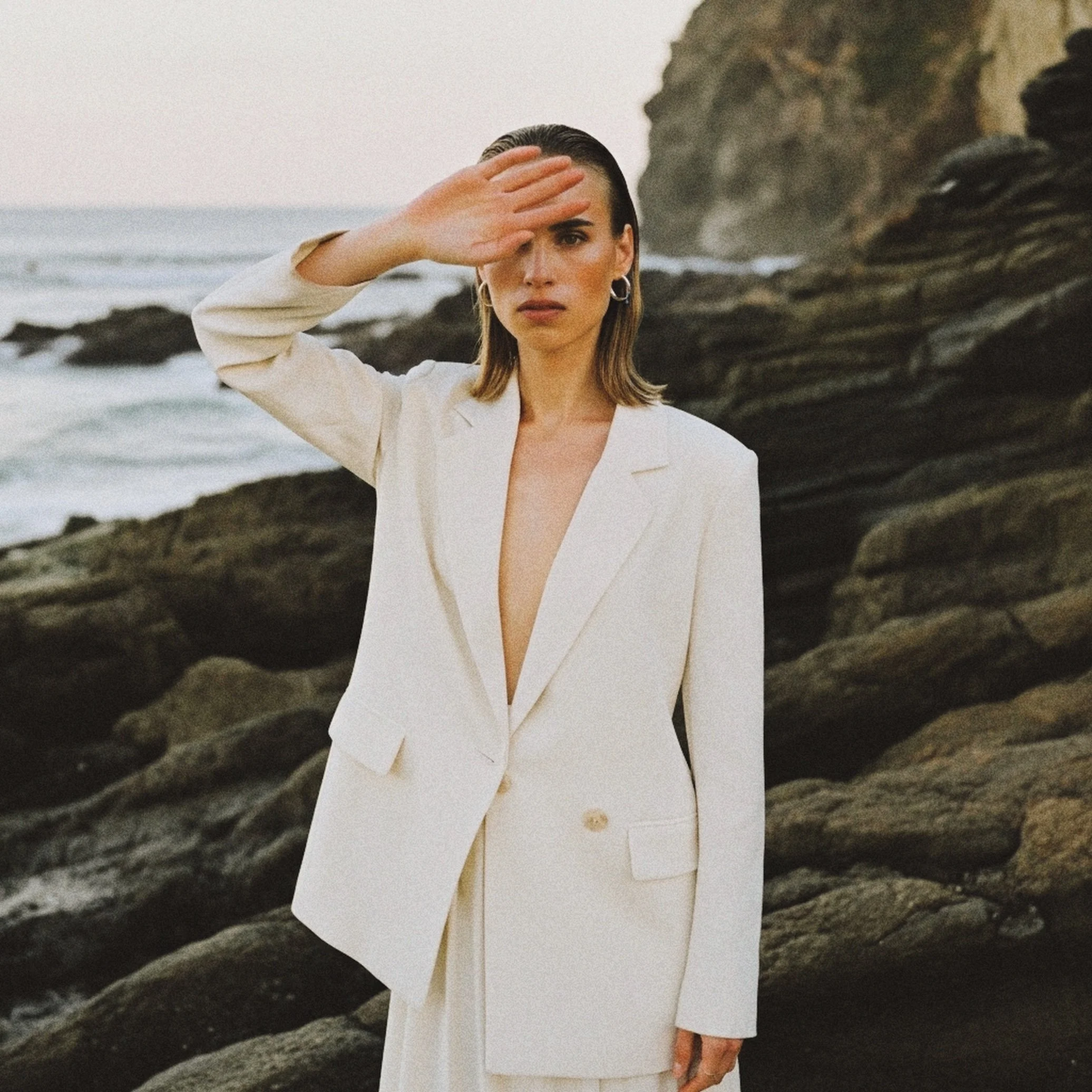 A woman in a cream-colored suit with a deep neckline standing on a rocky beach, shielding her eyes from the sun with her hand, with ocean waves and cliffs in the background.
