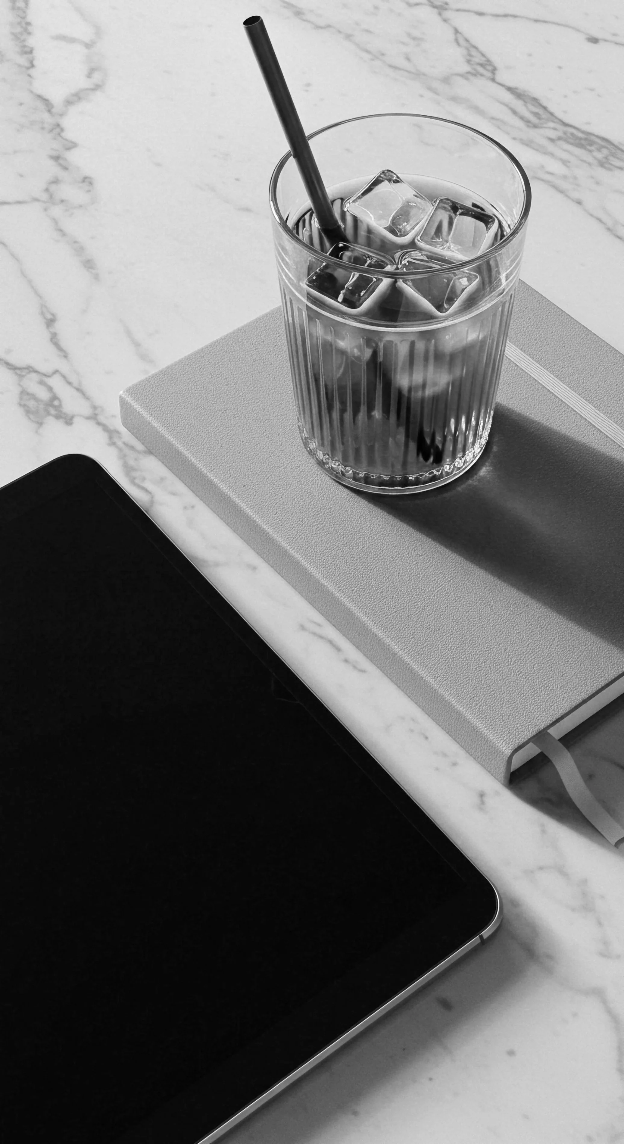 A glass of water with ice cubes and a straw, placed on top of a closed notebook on a marble surface, with part of a tablet visible in the foreground.
