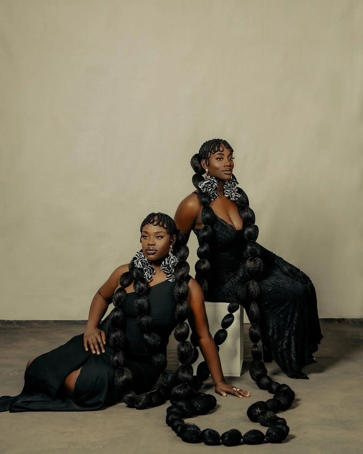 Two women with large, elaborate braided hairstyles and matching zebra-print hair scrunchies, dressed in black evening gowns, posing against a plain wall.