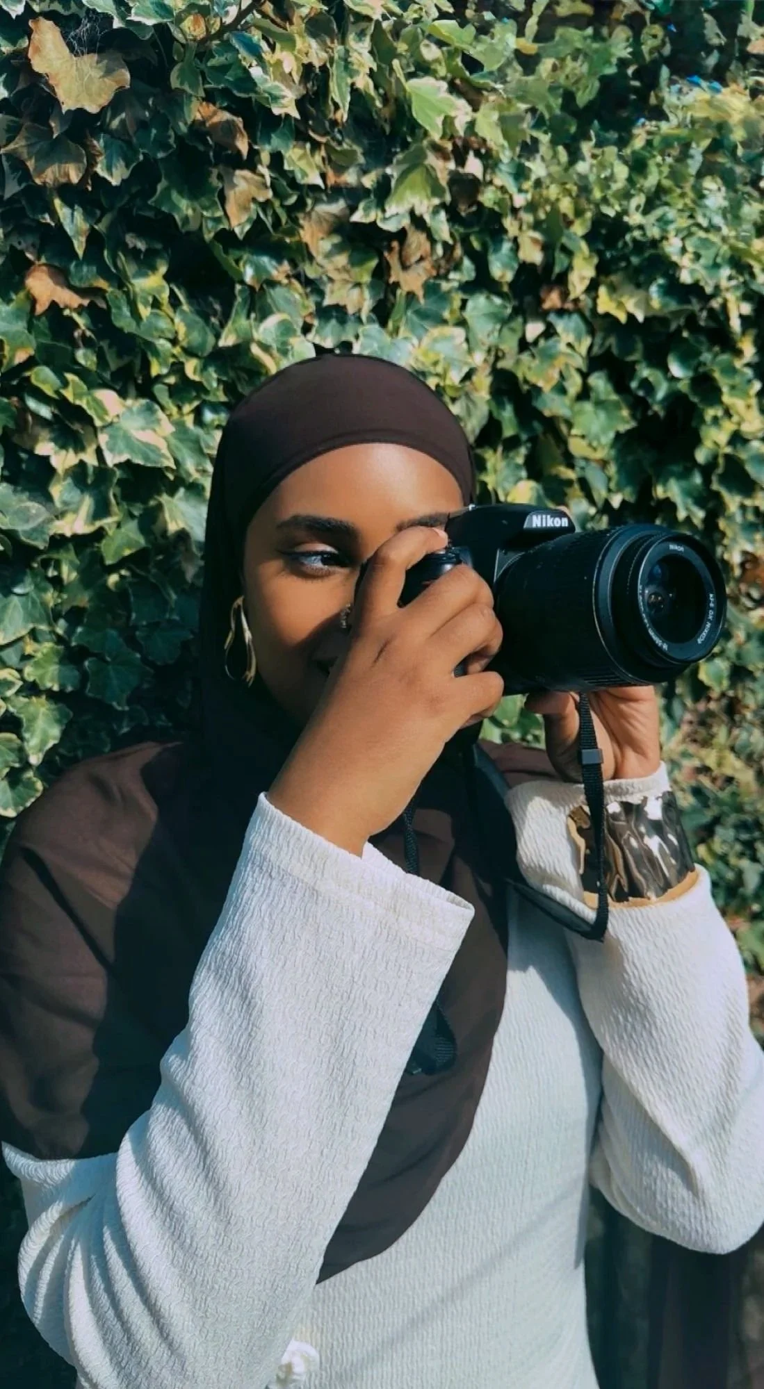 A woman wearing a brown hijab and white long-sleeve shirt, holding a Nikon DSLR camera to her eye, standing in front of a leafy green background.