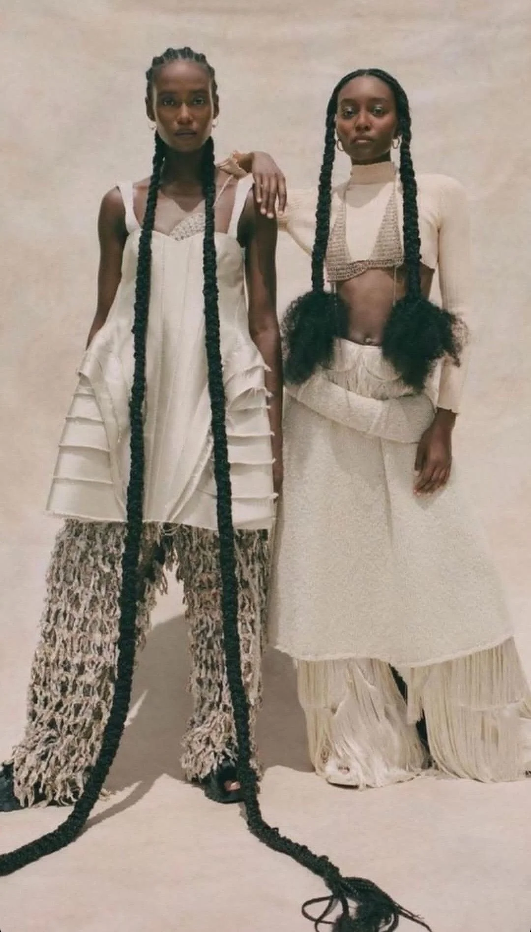 Two women standing against a beige wall, wearing fashion-forward, textured outfits with long braids and confident expressions.