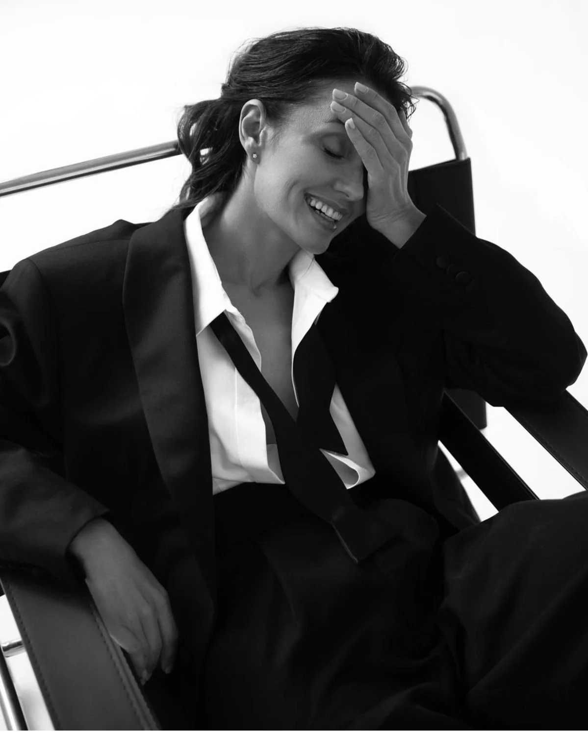 A woman in professional attire, sitting in a chair, smiling with her hand on her forehead.