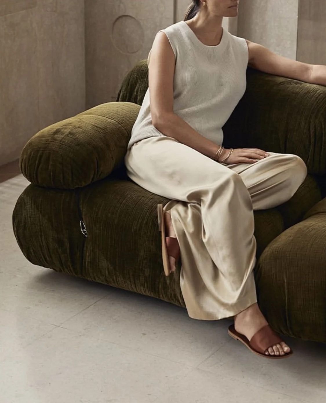 A woman sits on a green velvet sofa in a neutral-colored room, wearing a sleeveless white top, beige satin wide-leg pants, and brown open-toe sandals.