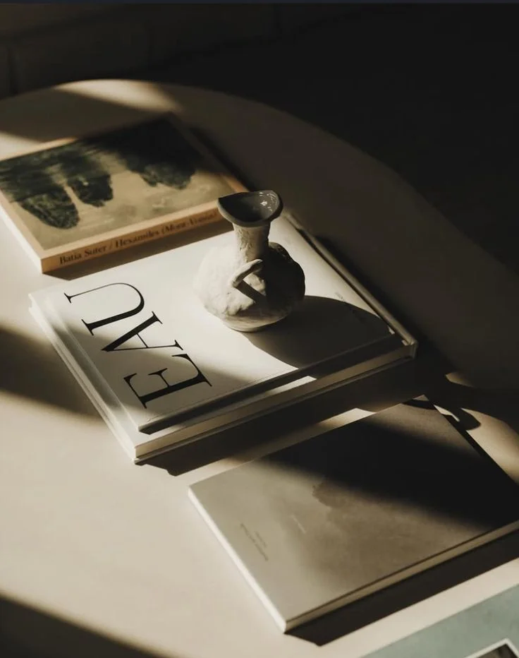Close-up of a stack of books with sunlight casting shadows on them, a small sculpture of a goose sitting on top of the book titled "EAT".