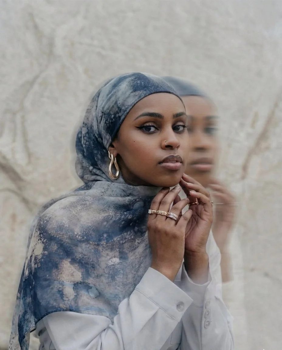A woman with dark skin and brown eyes wearing a blue and gray patterned headscarf and large hoop earrings, standing against a textured beige wall, with her reflection visible in a mirror behind her.