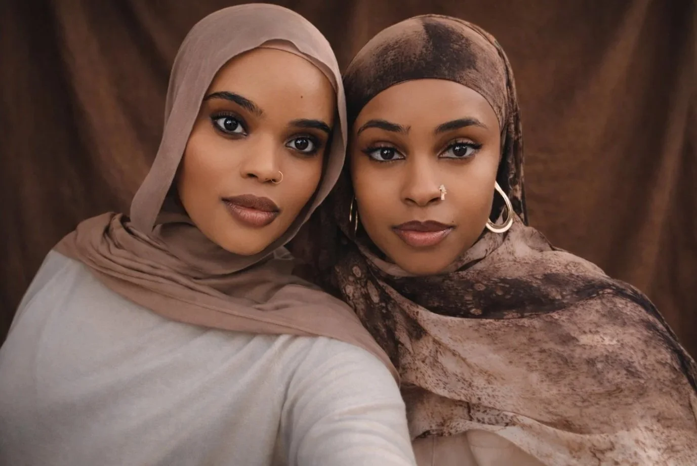 Two women wearing headscarves, one in beige and the other in brown, posing together against a brown fabric background.