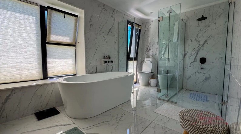 Baño moderno con bañera independiente, ducha de vidrio, inodoro, ventana con persianas, suelo y paredes de mármol blanco, y un recipiente de mimbre con almohadón.