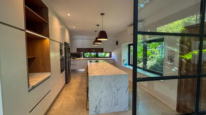 Cocina moderna con encimera de mármol y armarios blancos, ventanales grandes con vistas a un jardín verde