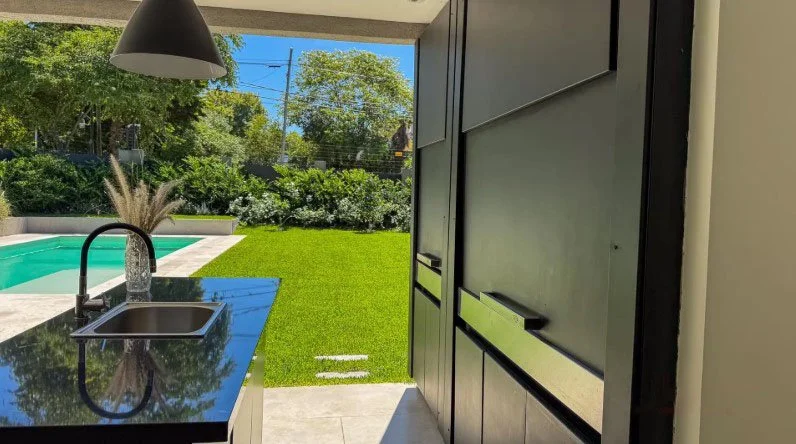Vista desde una cocina con encimera de piedra, fregadero y grifo, hacia un jardín con césped y piscina, con árboles y un cielo despejado.