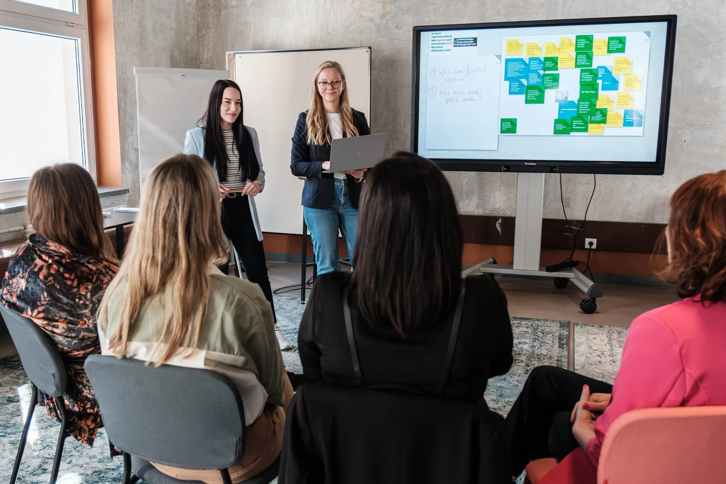 Two colleagues explaining notes and process flows on a whiteboard to others during a team meeting