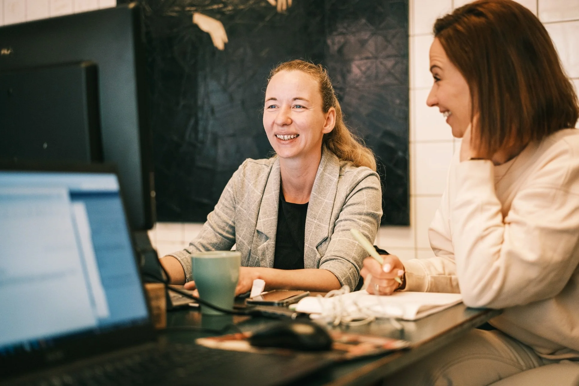 Two colleagues collaborating at a desk, smiling while reviewing work on a computer during a meeting