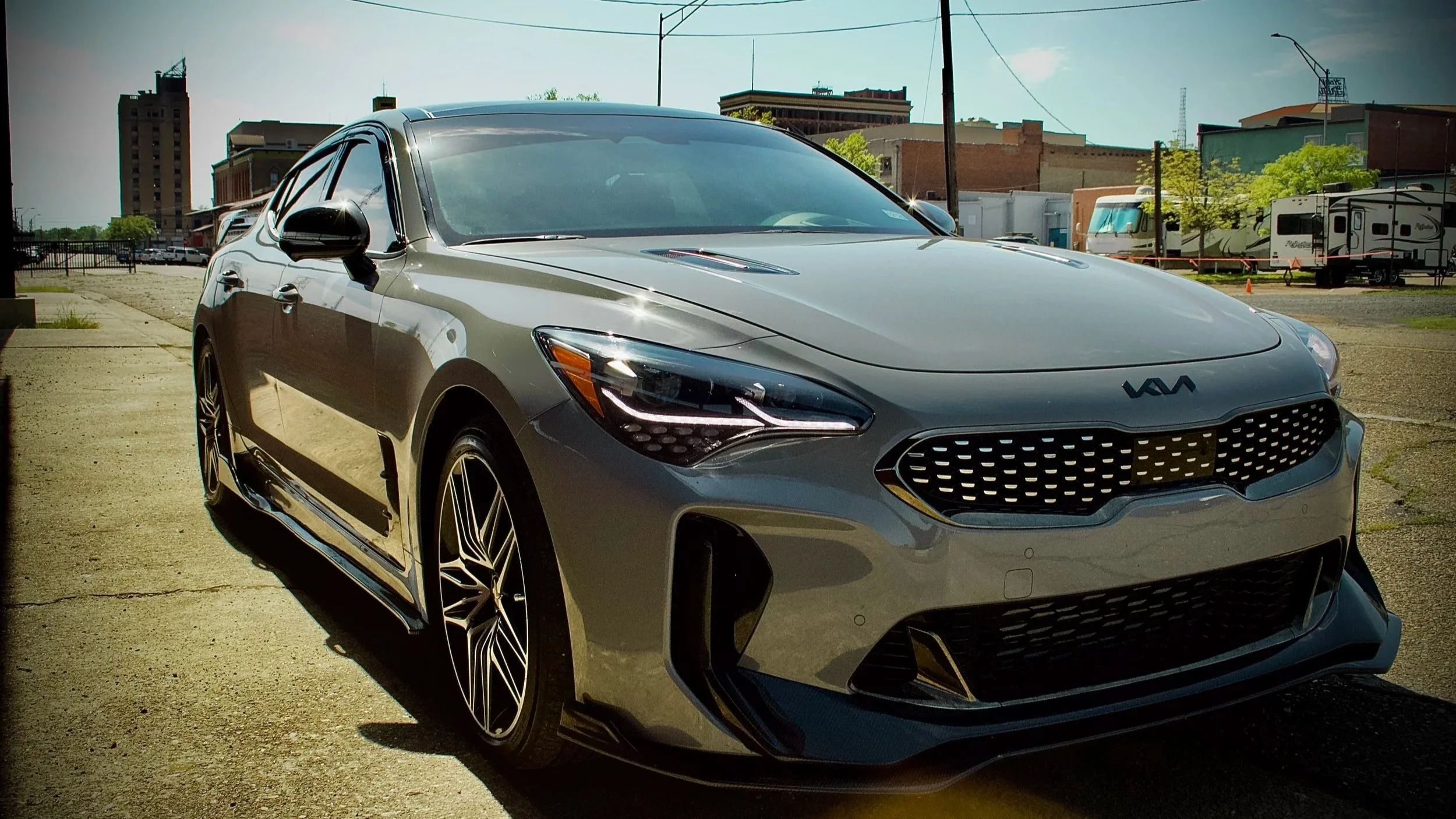 Silver Kia Stinger sports sedan parked on a dirt lot with industrial buildings, RVs, and power lines in the background.