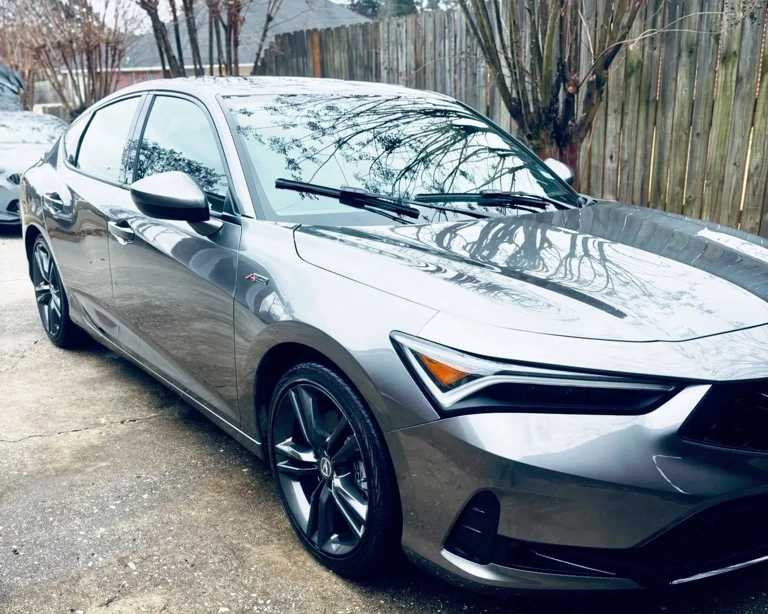 A silver modern sedan car parked on a dirt surface, with a wooden fence and trees in the background.