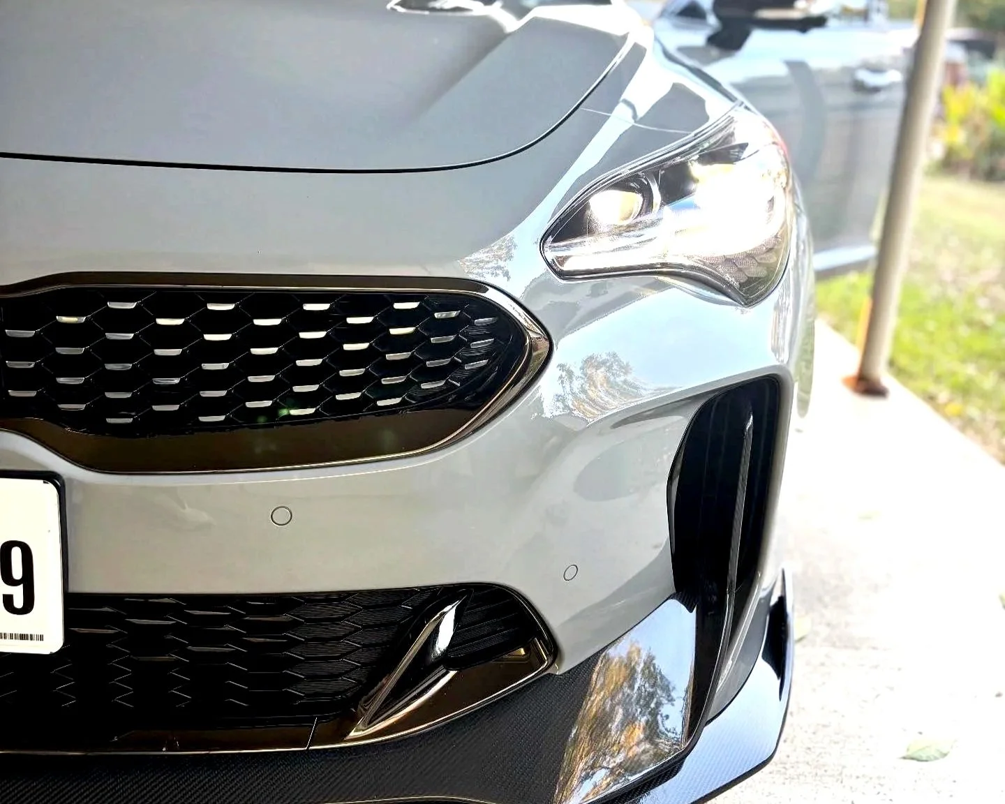 Close-up of a silver luxury car's front end, showing a black mesh grille, headlight, part of the bumper, and a front splitter.