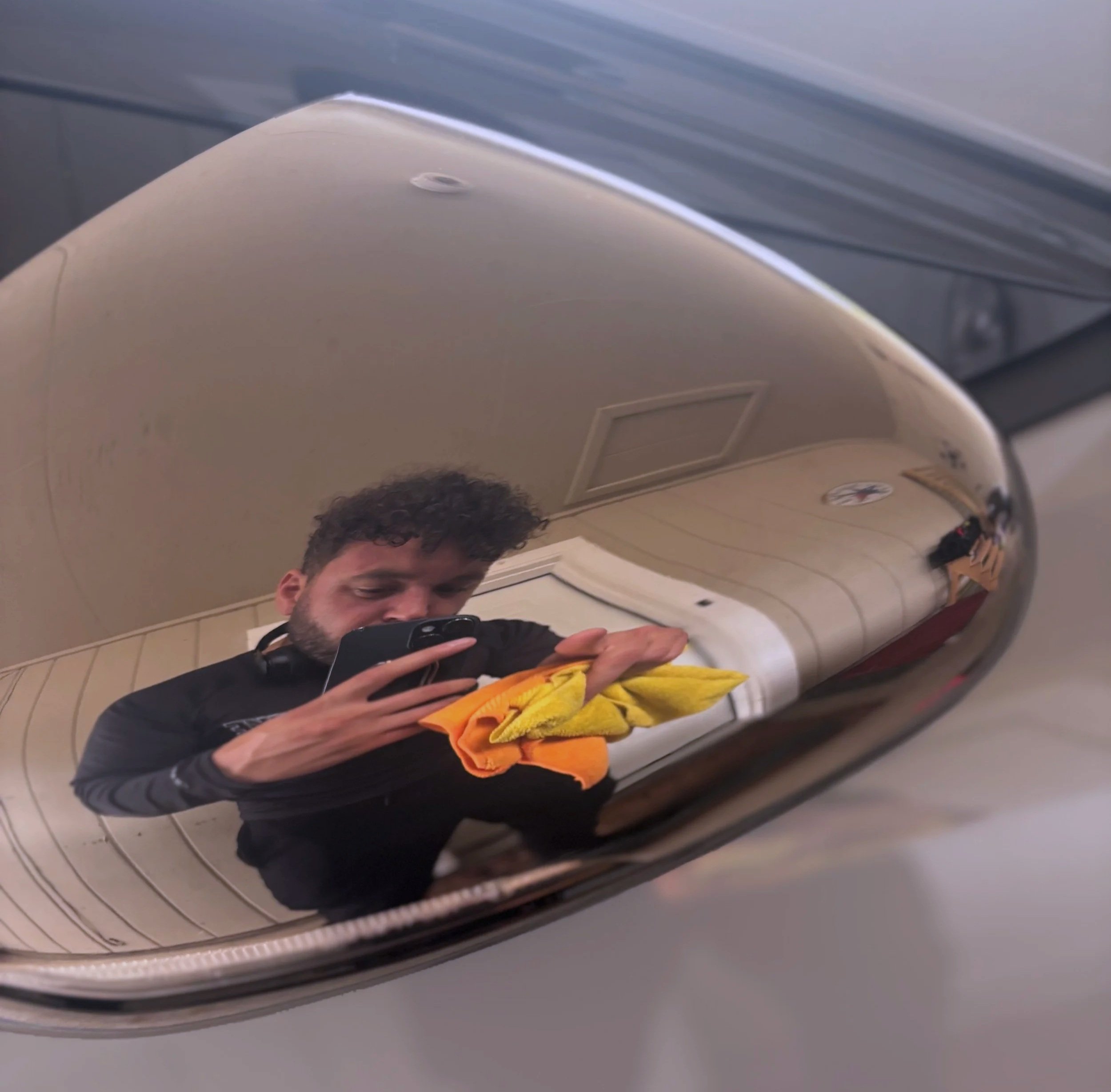 Reflection of a man taking a selfie with his phone in a shiny, curved surface, showing a room with beige walls and wooden paneling, a door, and a Pittsburgh Steelers logo.