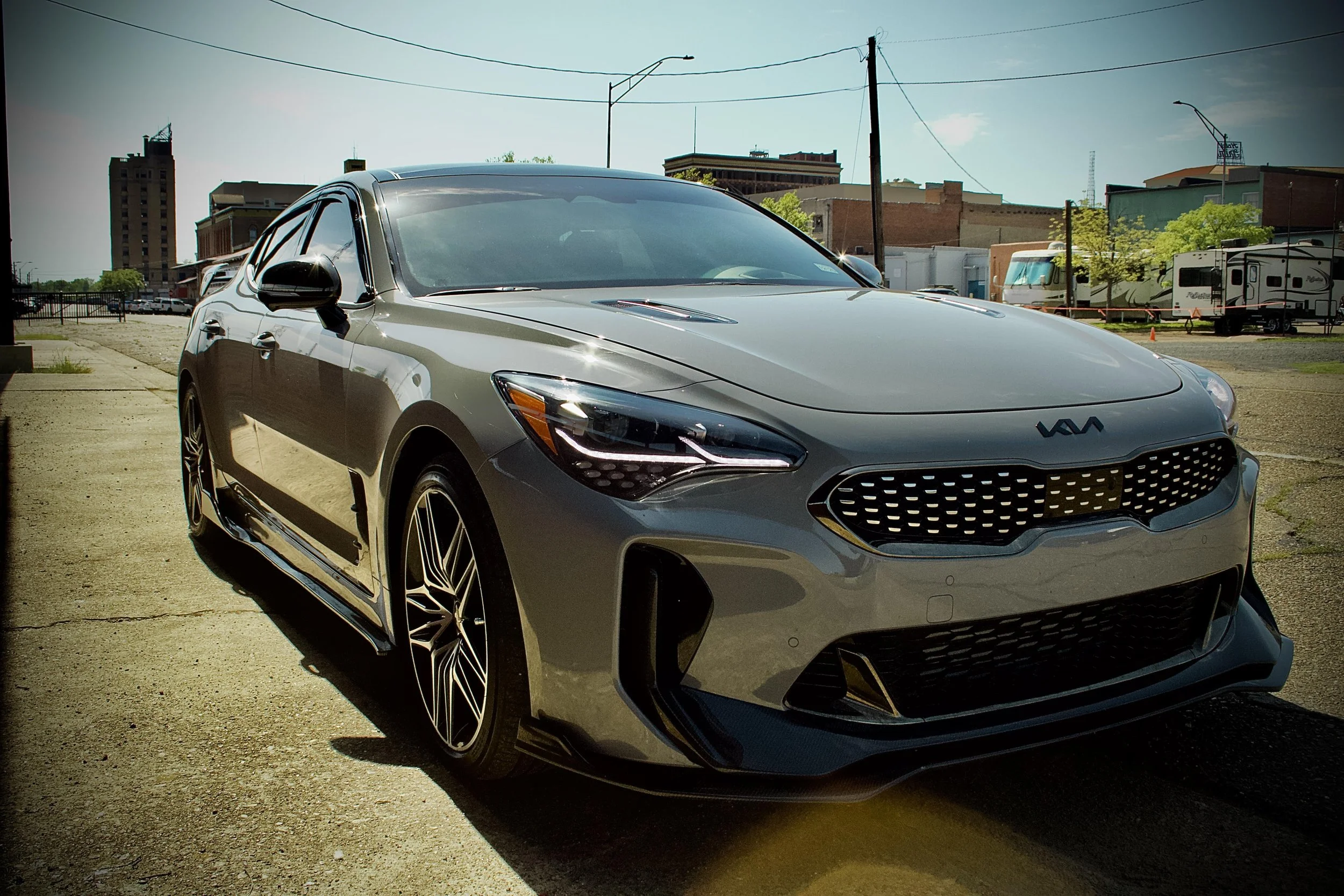 Gray Kia sedan parked on a city street with buildings and RVs in the background on a sunny day.