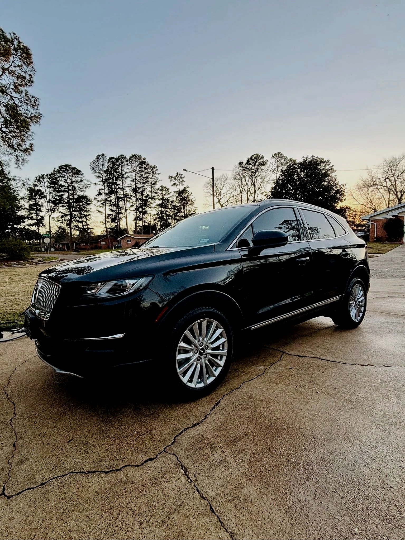 Black SUV parked on a cracked concrete driveway with trees and houses in the background during sunset.