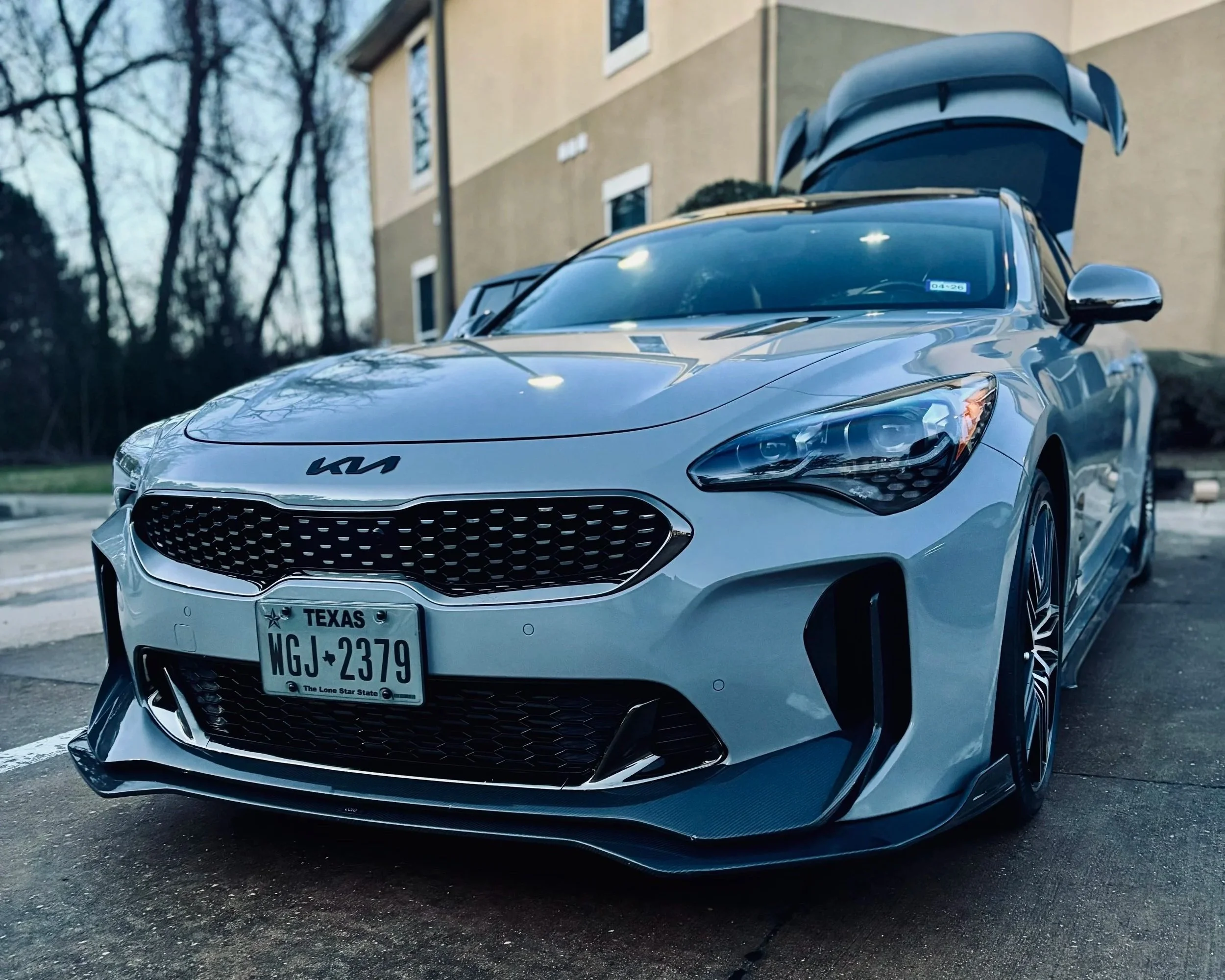 A silver Kia car with a Texas license plate, parked on a street with a building and trees in the background. The hood is slightly open.