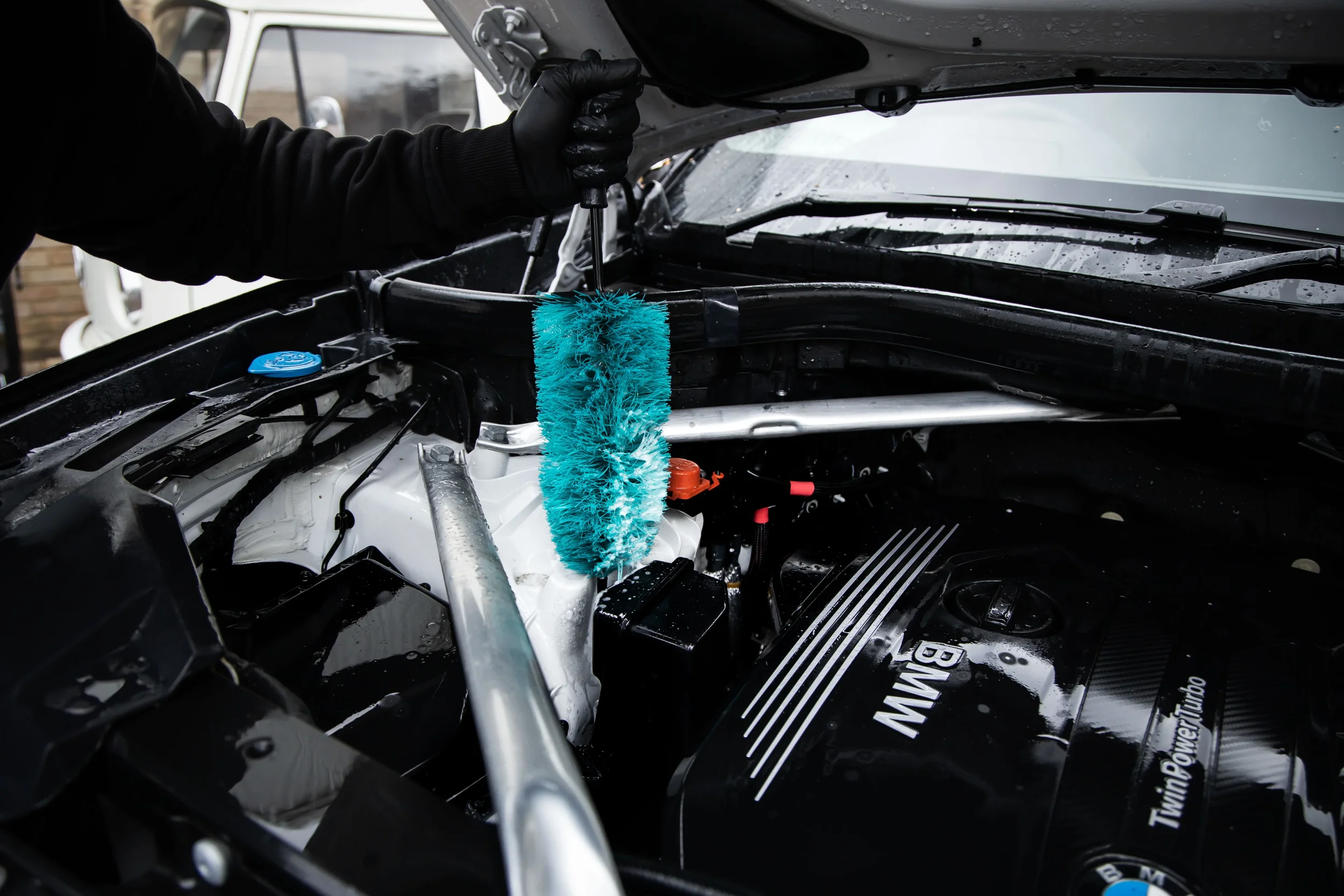 A person wearing black gloves holds a brush while cleaning the engine of a BMW vehicle. The engine features a black cover with the BMW logo and the word 'TwinPower Turbo.' Water droplets are visible on the windshield.