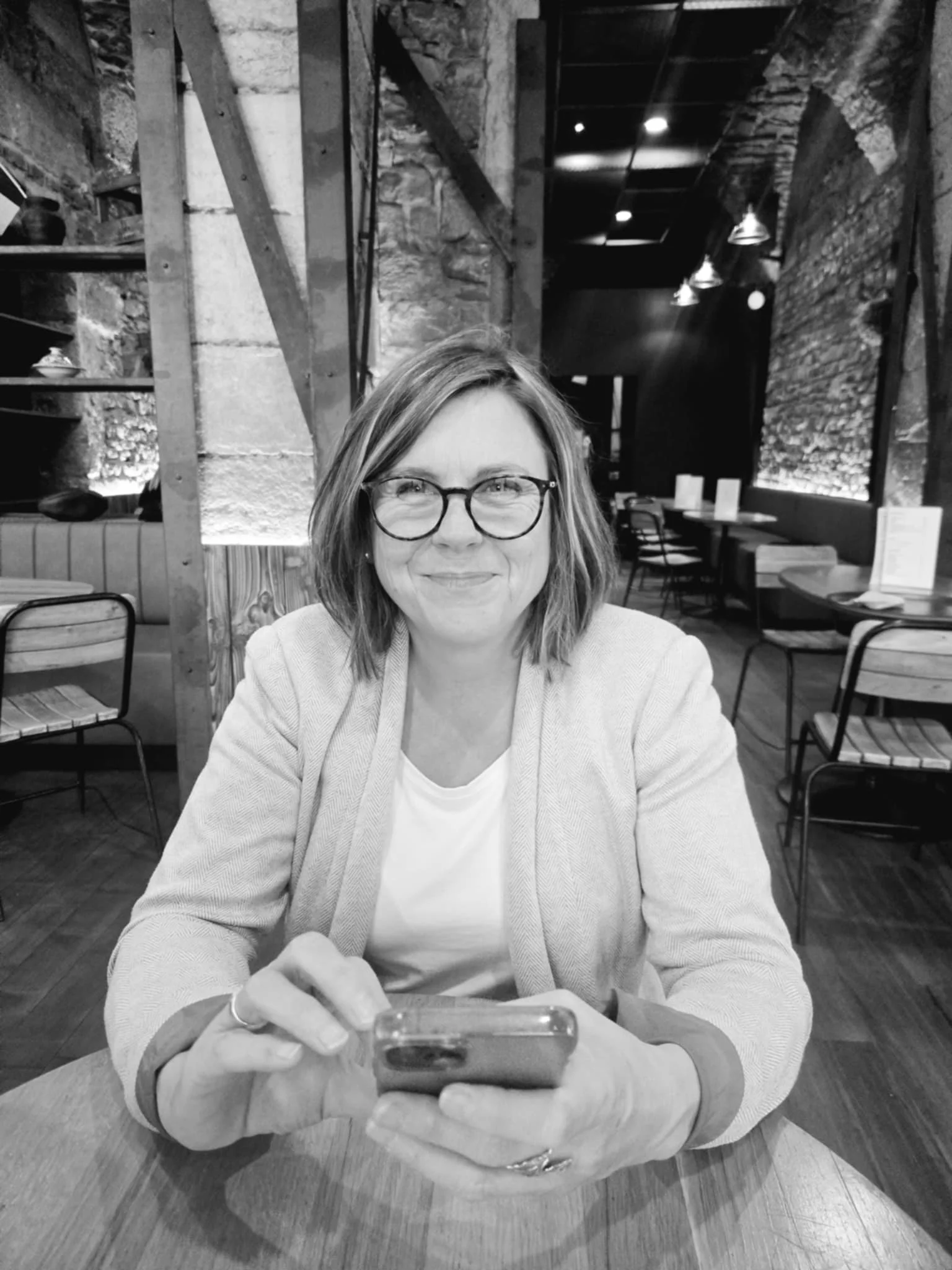 Femme souriante portant des lunettes, assise à une table dans un restaurant ou café, tenant un téléphone portable, décor en pierre et bois, ambiance chaleureuse et moderne.