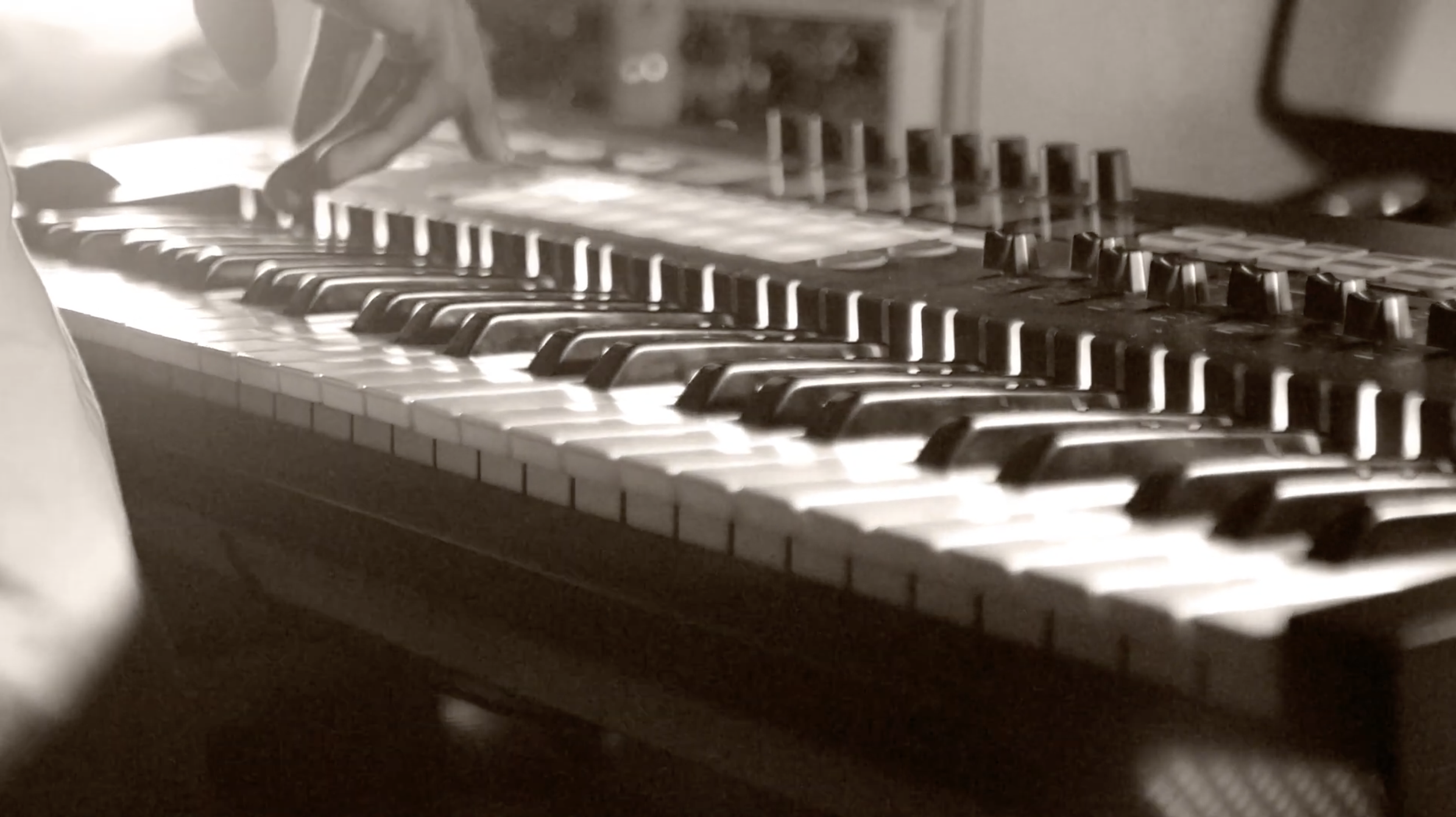 Close-up of a keyboard synthesizer with black and white keys and various knobs on top, seen in black and white. Video created by graphic design students Kenzie Coates and Katherine Lancaster.