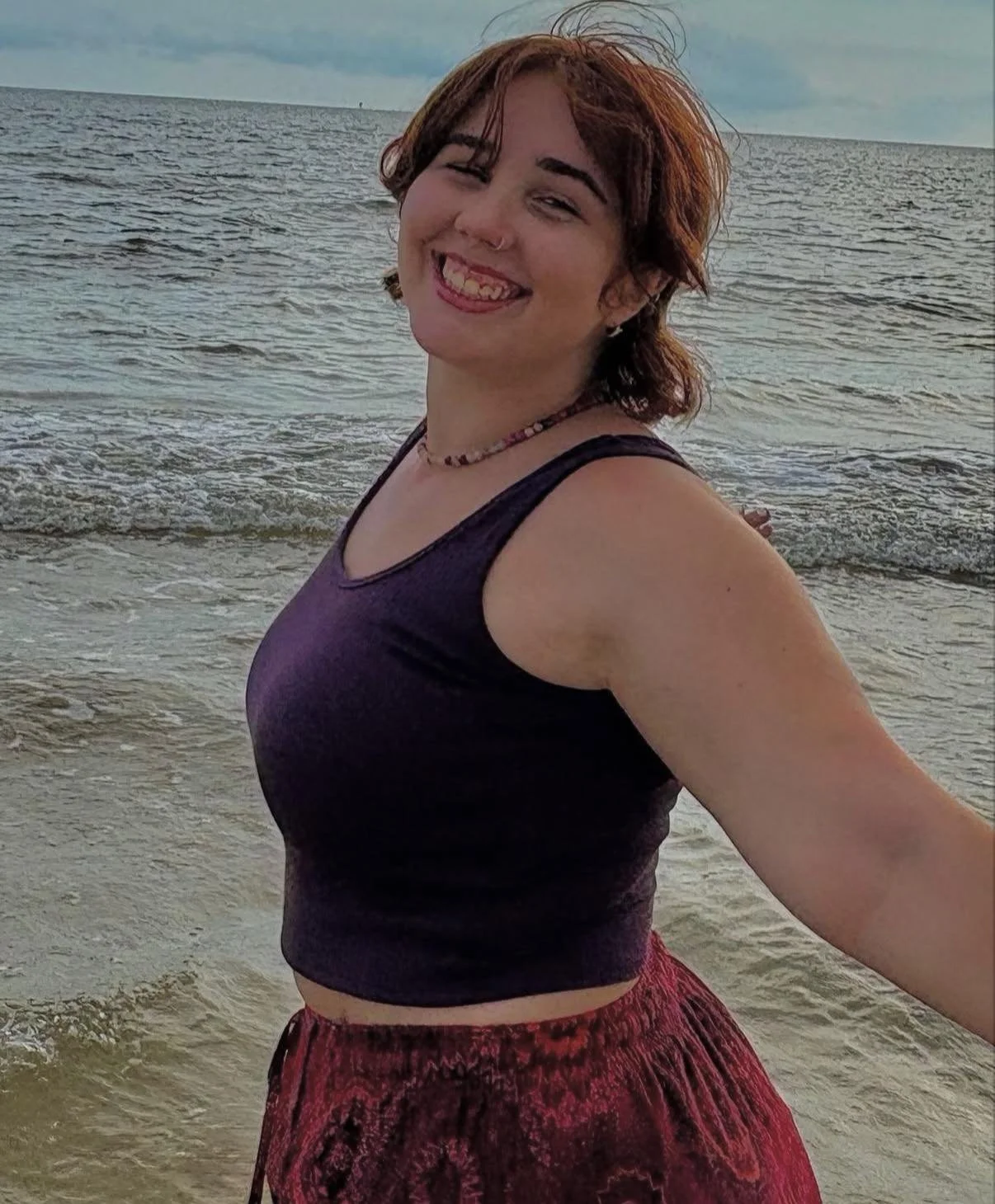 Casual image of graphic design student Kenzie Coates at the beach, wearing a purple sleeveless top and purple patterned pants, smiling at the camera.