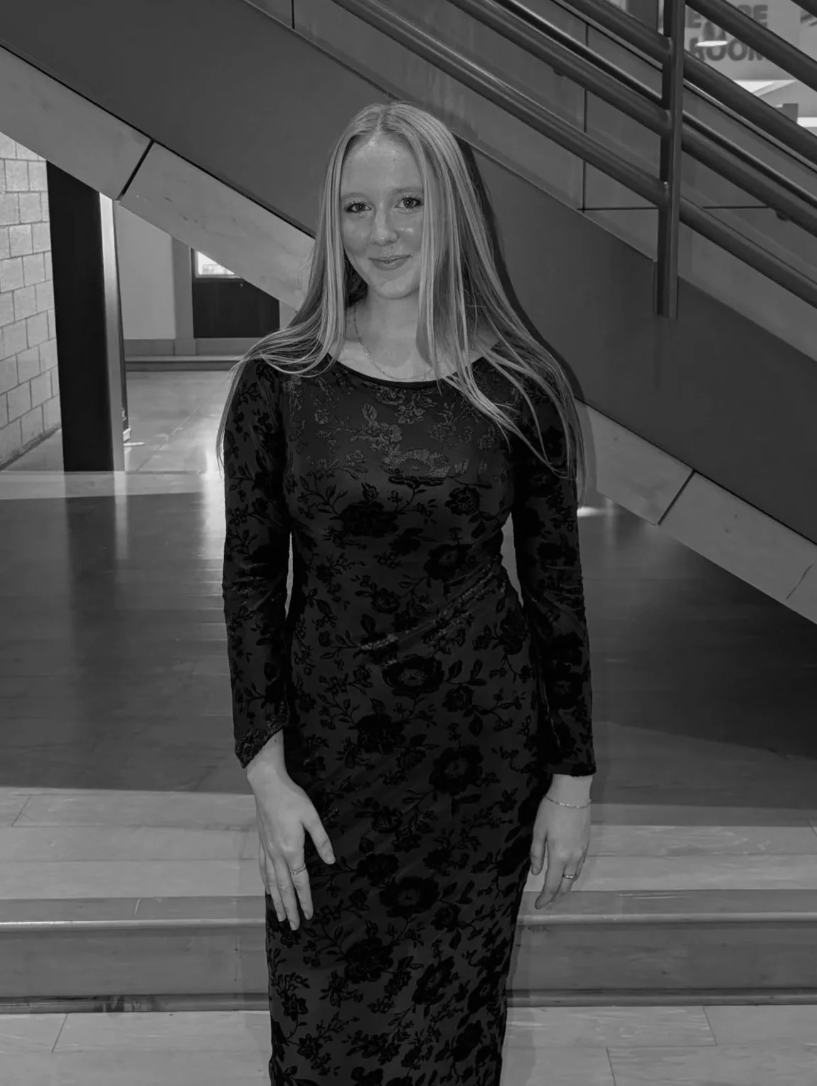 A young woman with long light hair, wearing a long-sleeved dark floral dress, smiling and standing indoors near wooden stairs and a metal railing.