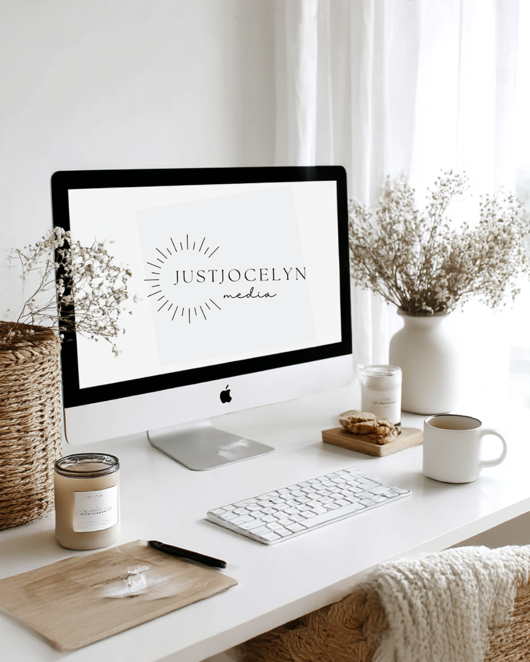 Minimalist workspace with a desktop computer displaying "Just Jocelyn Media" logo, surrounded by white flowers, a candle, a mug, cookies, and a pen on a wooden board.